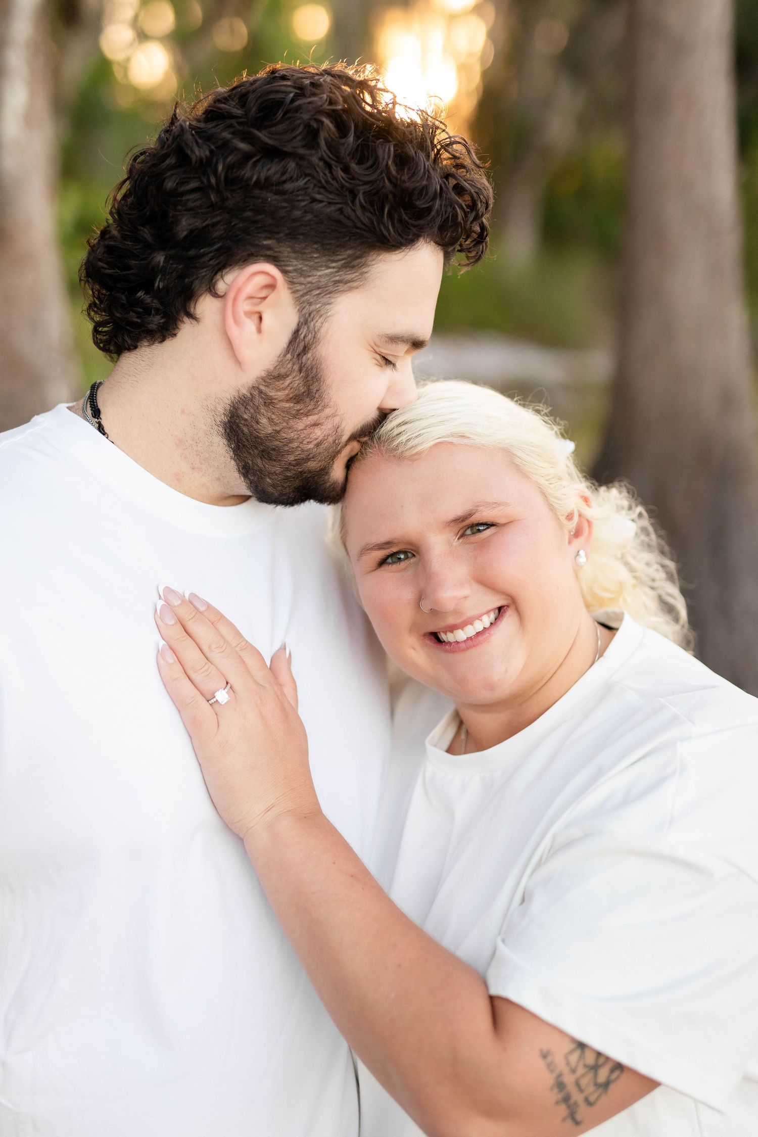 Man kissing woman's forehead as she smiles and shows her engagement ring, both wearing white shirts outdoors.