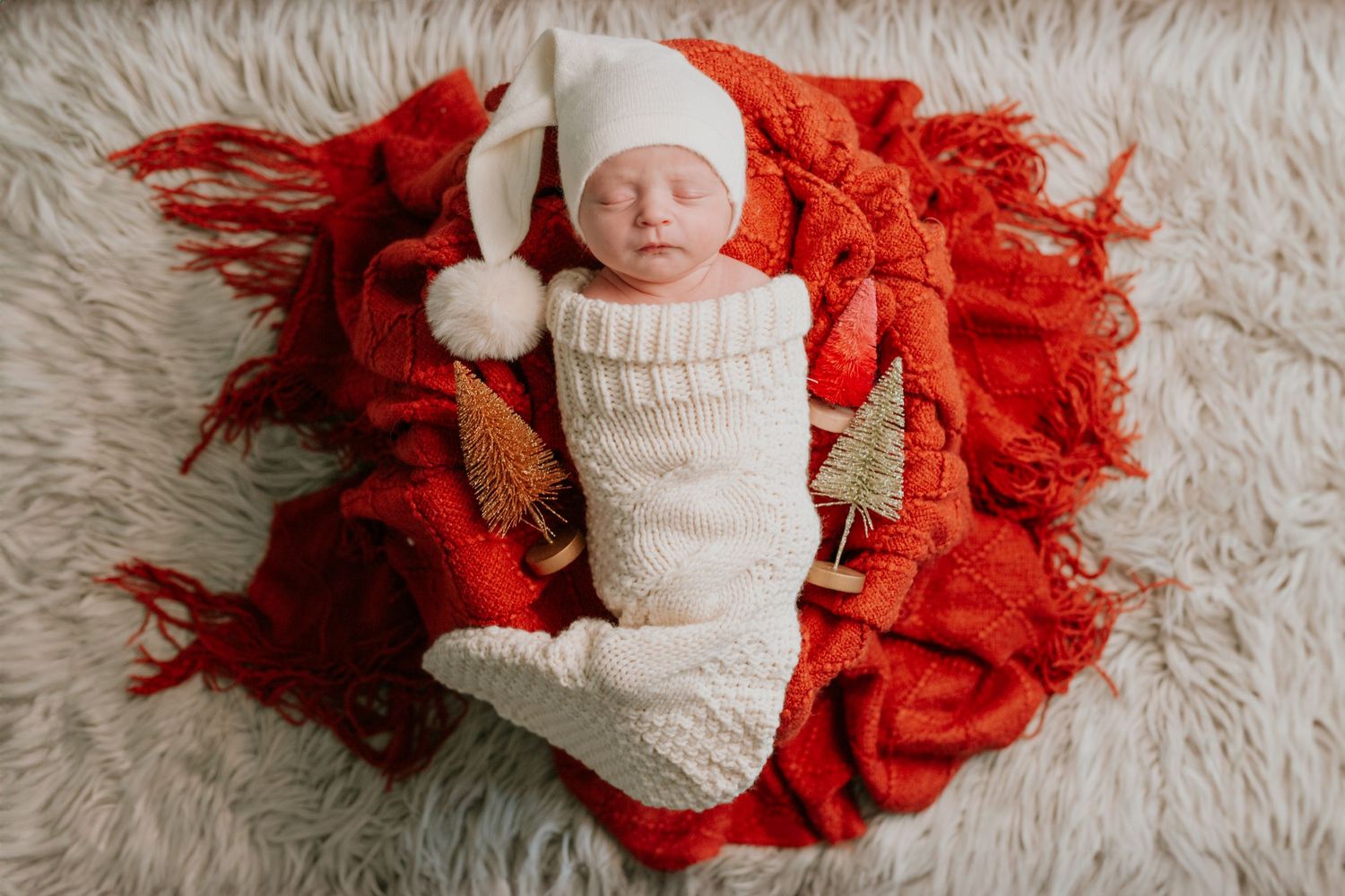 Newborn baby in cream knit Santa outfit and hat posed on red blanket for Christmas photo.