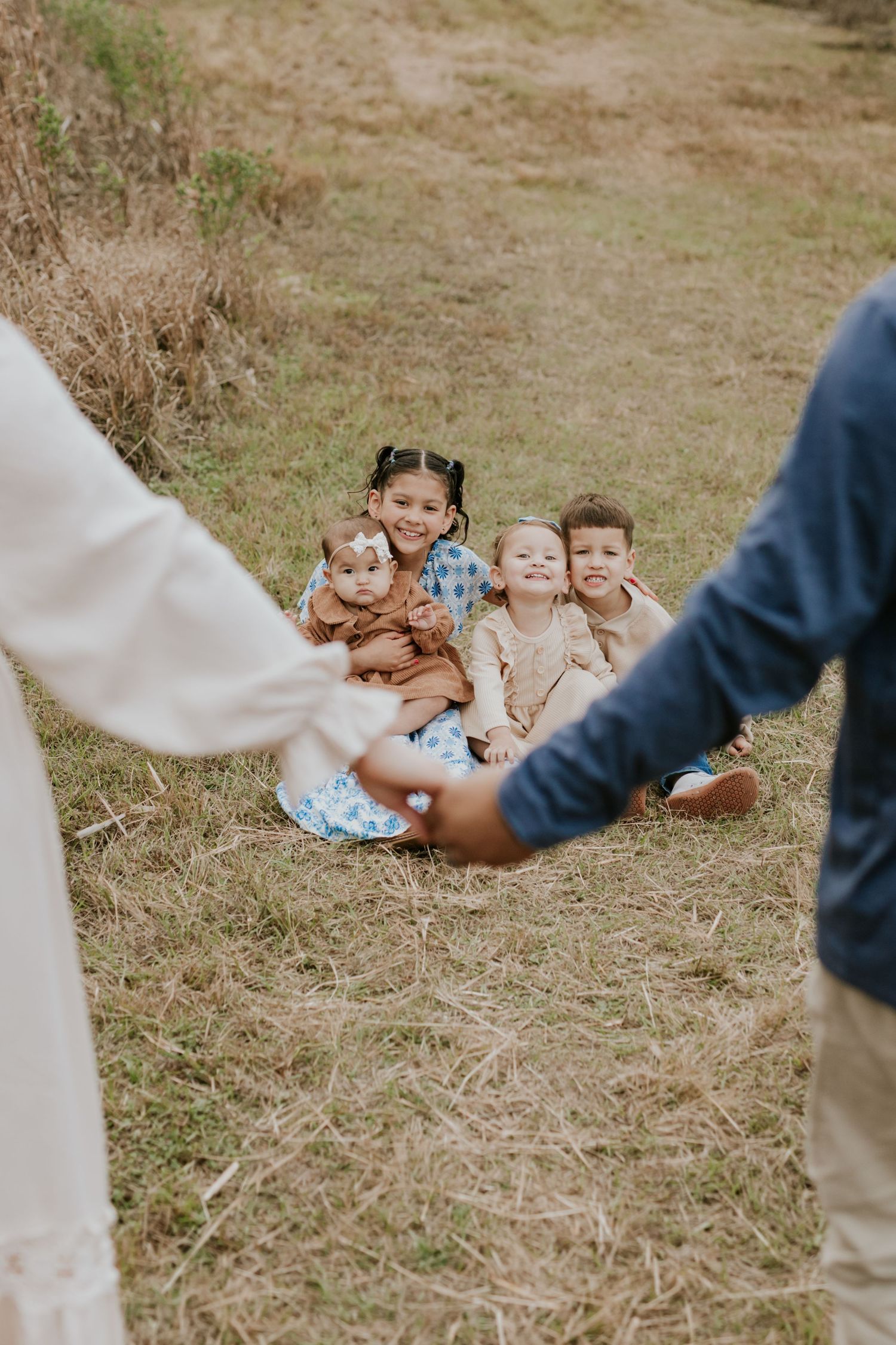 Four children sitting in grass while parents hold hands in foreground.