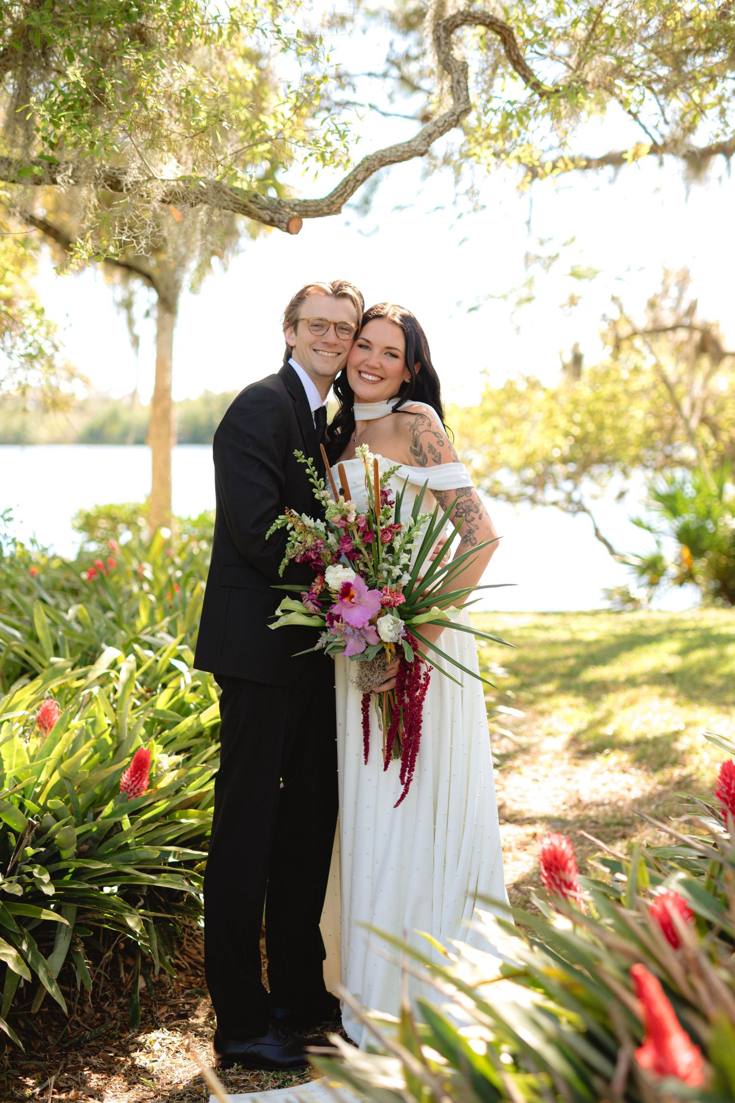 Newlywed couple embracing near a sunlit lakeside garden with lush tropical plants and a vibrant floral bouquet.