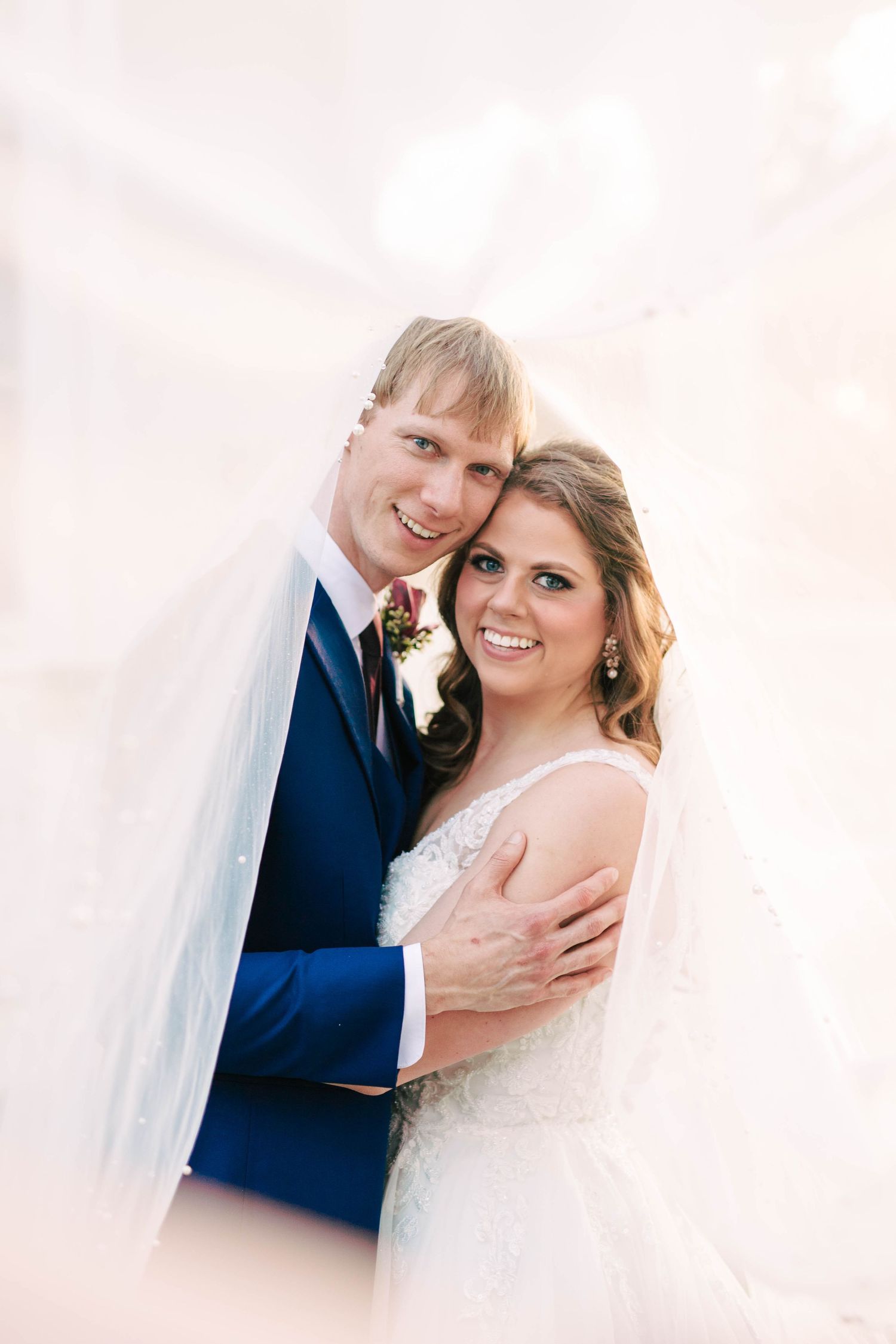 Groom embraces bride wrapped in a flowing veil, both smiling warmly at the camera on their wedding day.