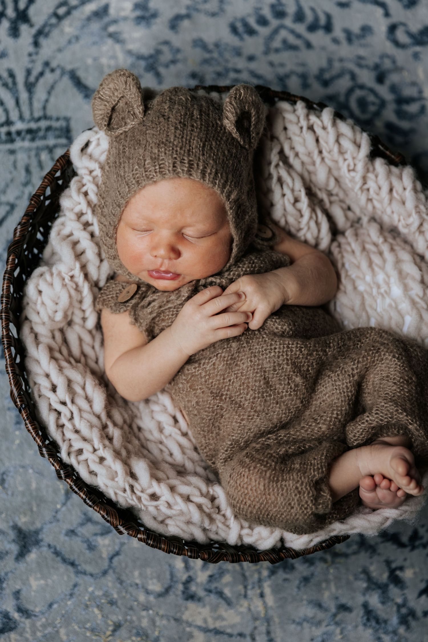 Sleeping newborn baby in knit bear outfit and hat resting in a wicker basket with cream blanket.