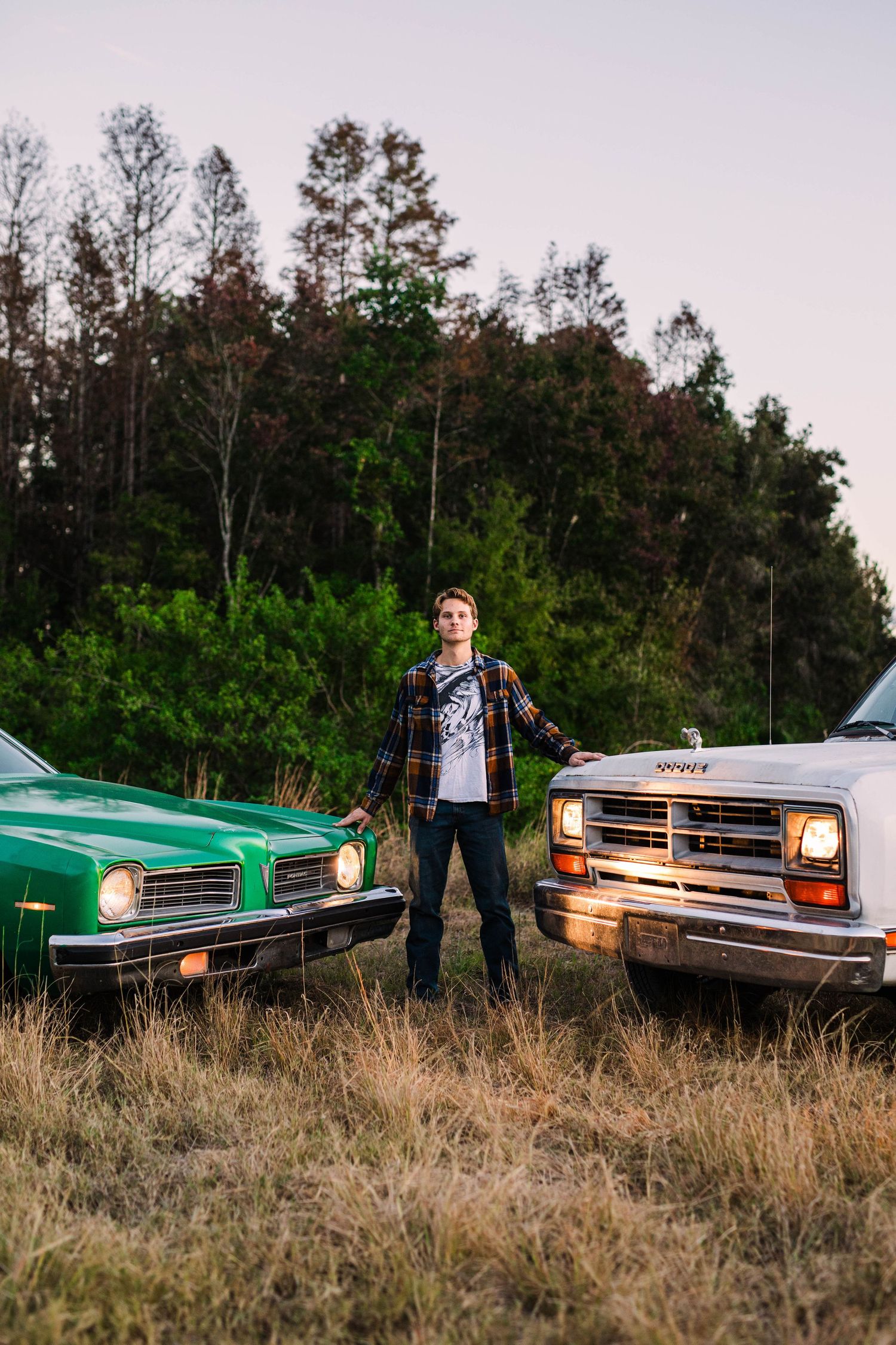 Young man standing between vintage green car and white truck in rural field at dusk.