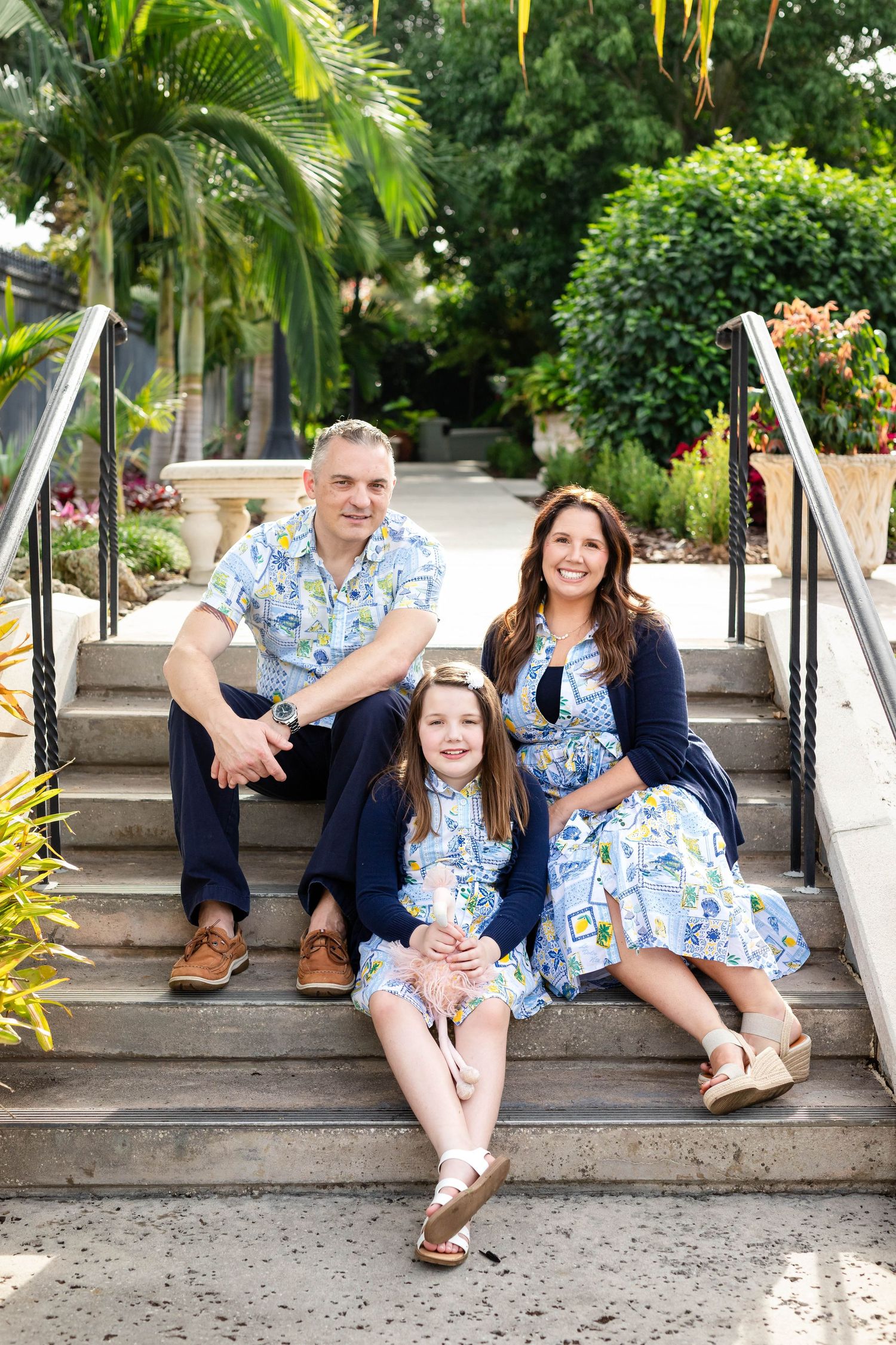 Cheerful family of three in coordinating blue outfits sits on outdoor steps with tropical greenery at Hollis Gardens.