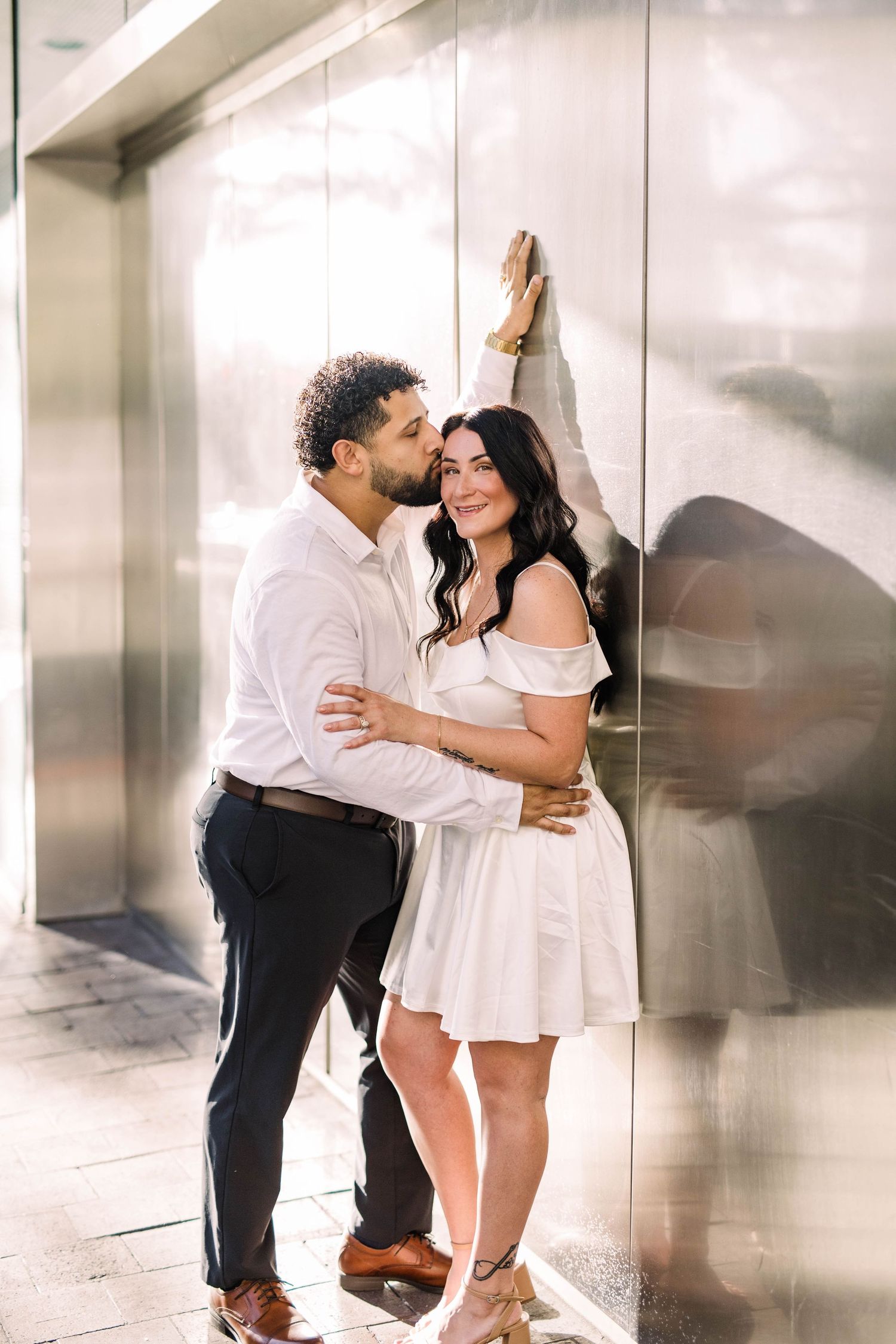 Couple posing against a reflective metallic wall, woman in white off-shoulder dress smiling joyfully in Tampa.
