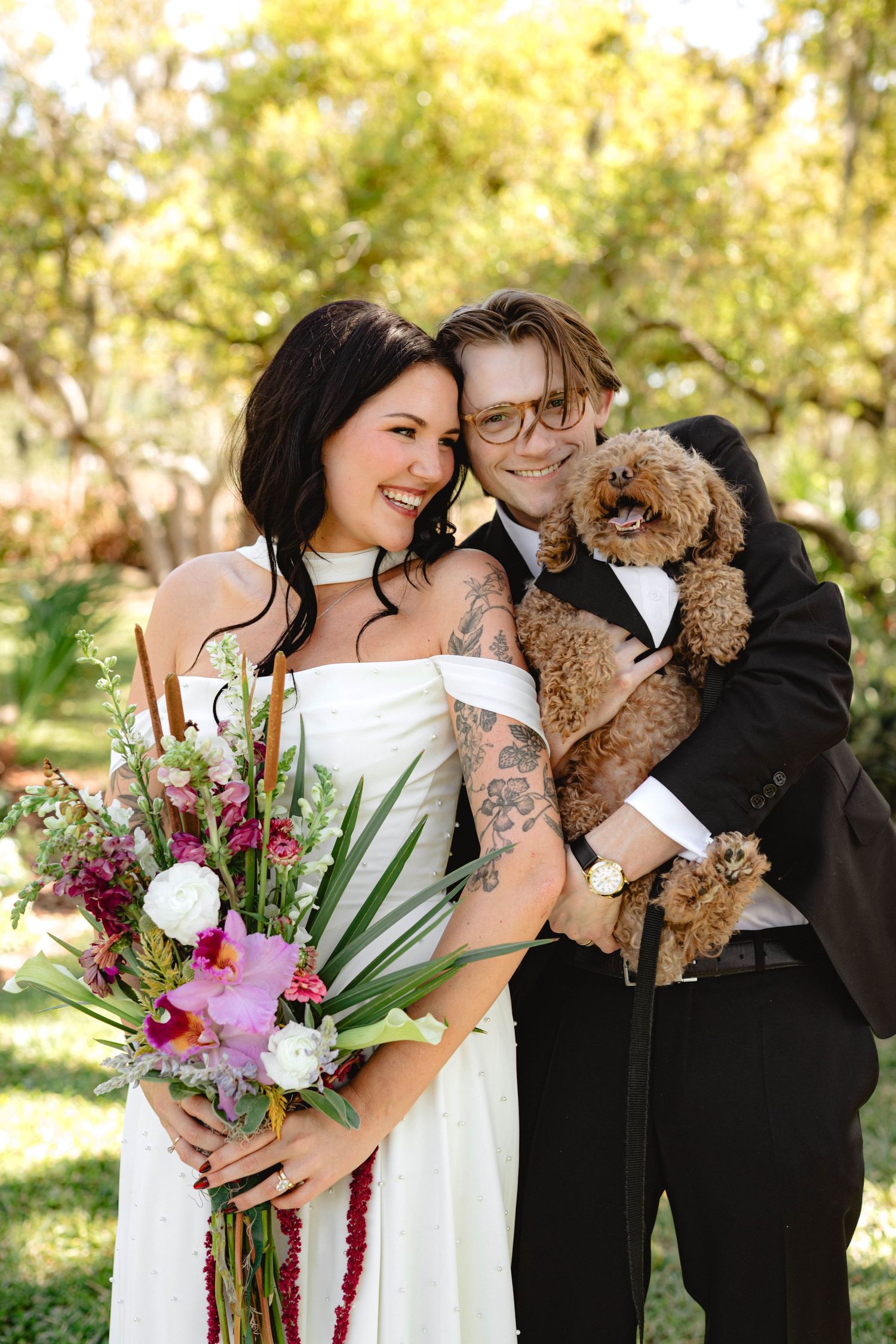 Bride and groom smiling together holding a colorful bouquet and fluffy brown dog in an outdoor garden setting.