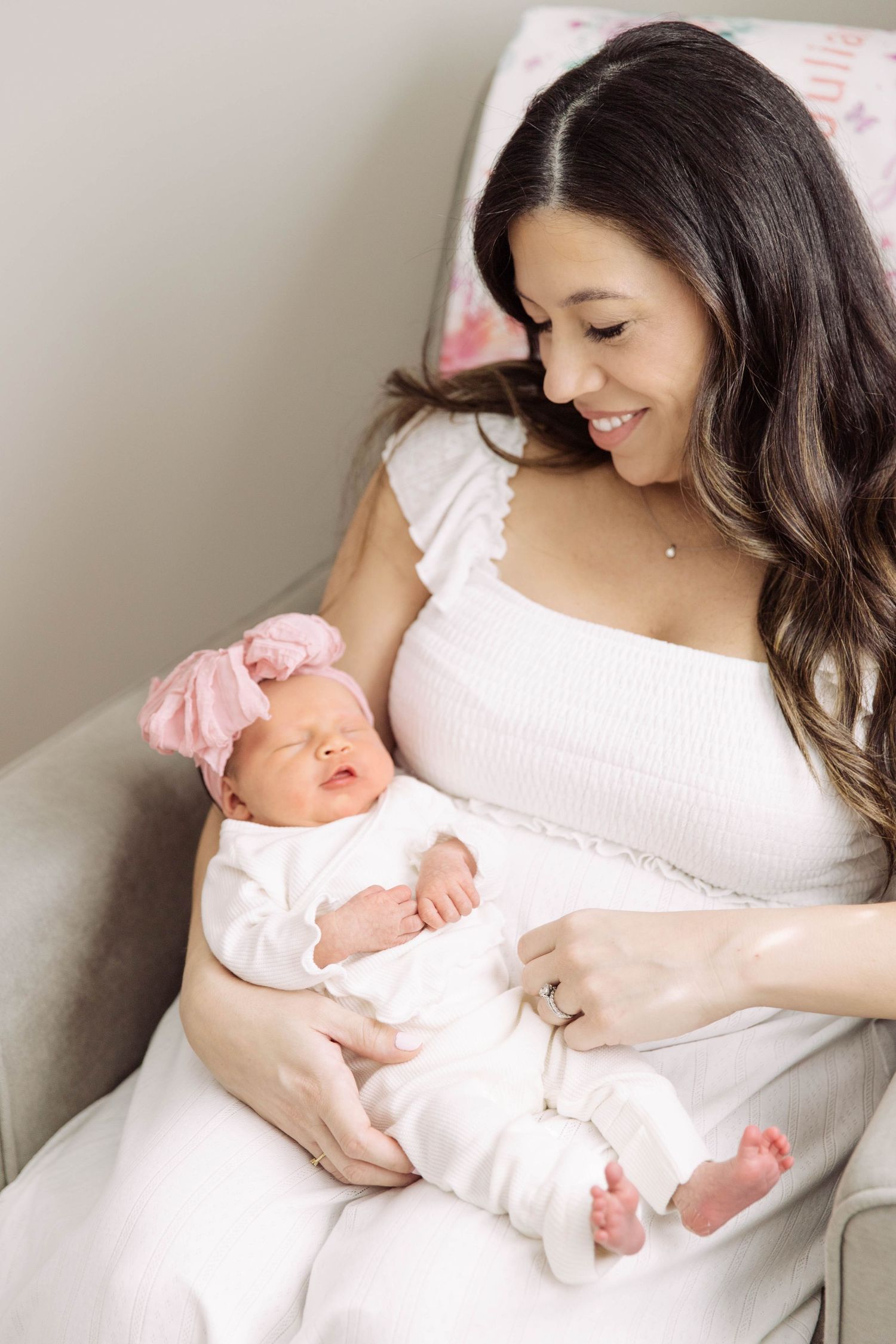 Smiling brunette mother in white dress gazes lovingly at her newborn with pink bow headband.