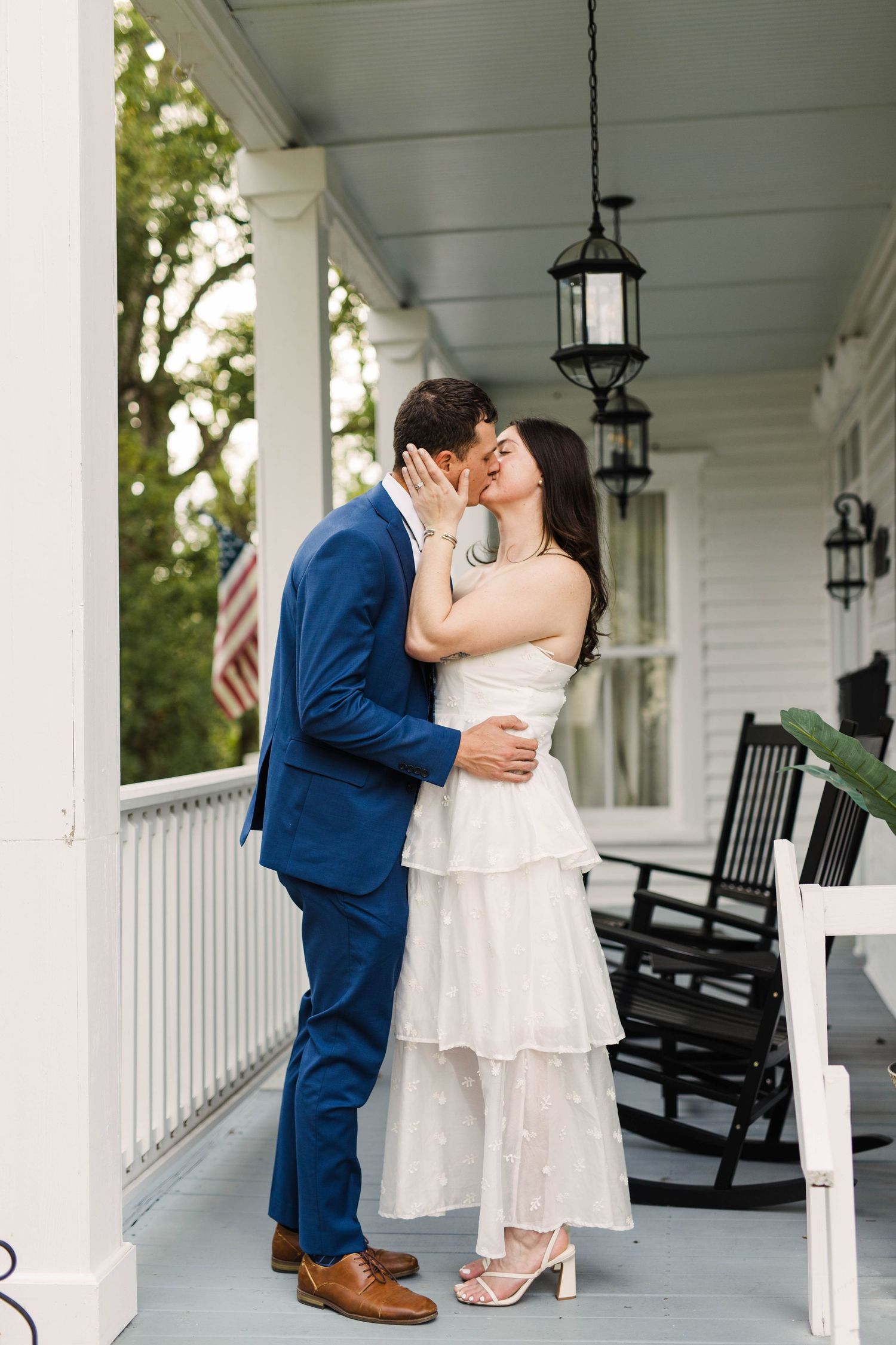 Couple shares a romantic kiss on a white porch with rocking chairs and hanging lanterns on their wedding day.