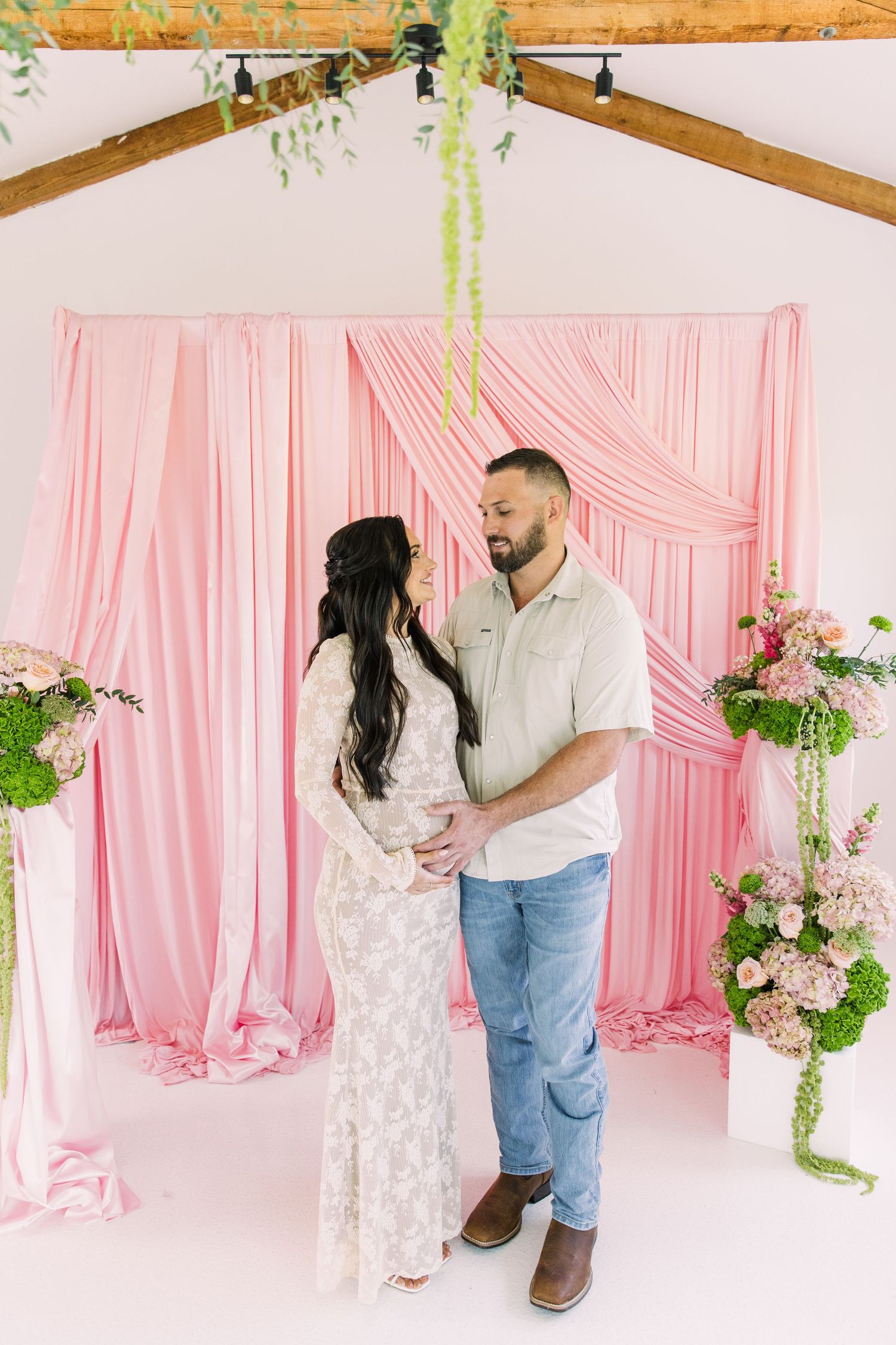 Pregnant woman in lace dress with husband standing before pink floral backdrop at the Colonial at Sunset..