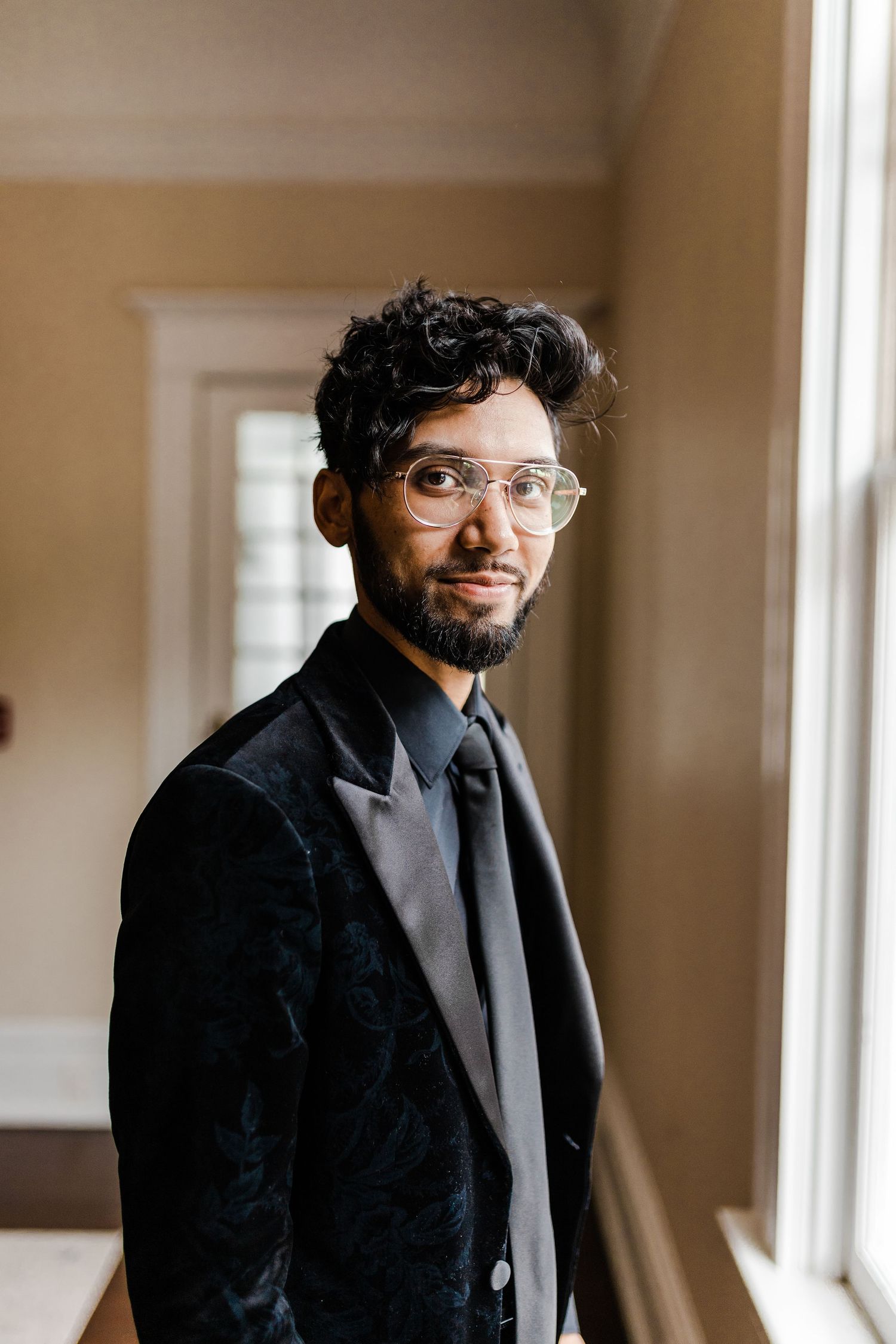 Stylish groom in black velvet blazer and glasses standing by window in soft indoor lighting at the Orlo.