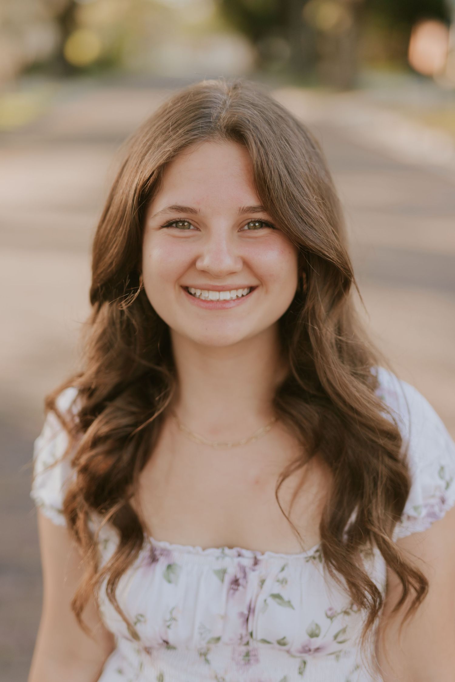 Young woman with long brown wavy hair smiling outdoors, wearing a white floral dress and delicate gold necklace.