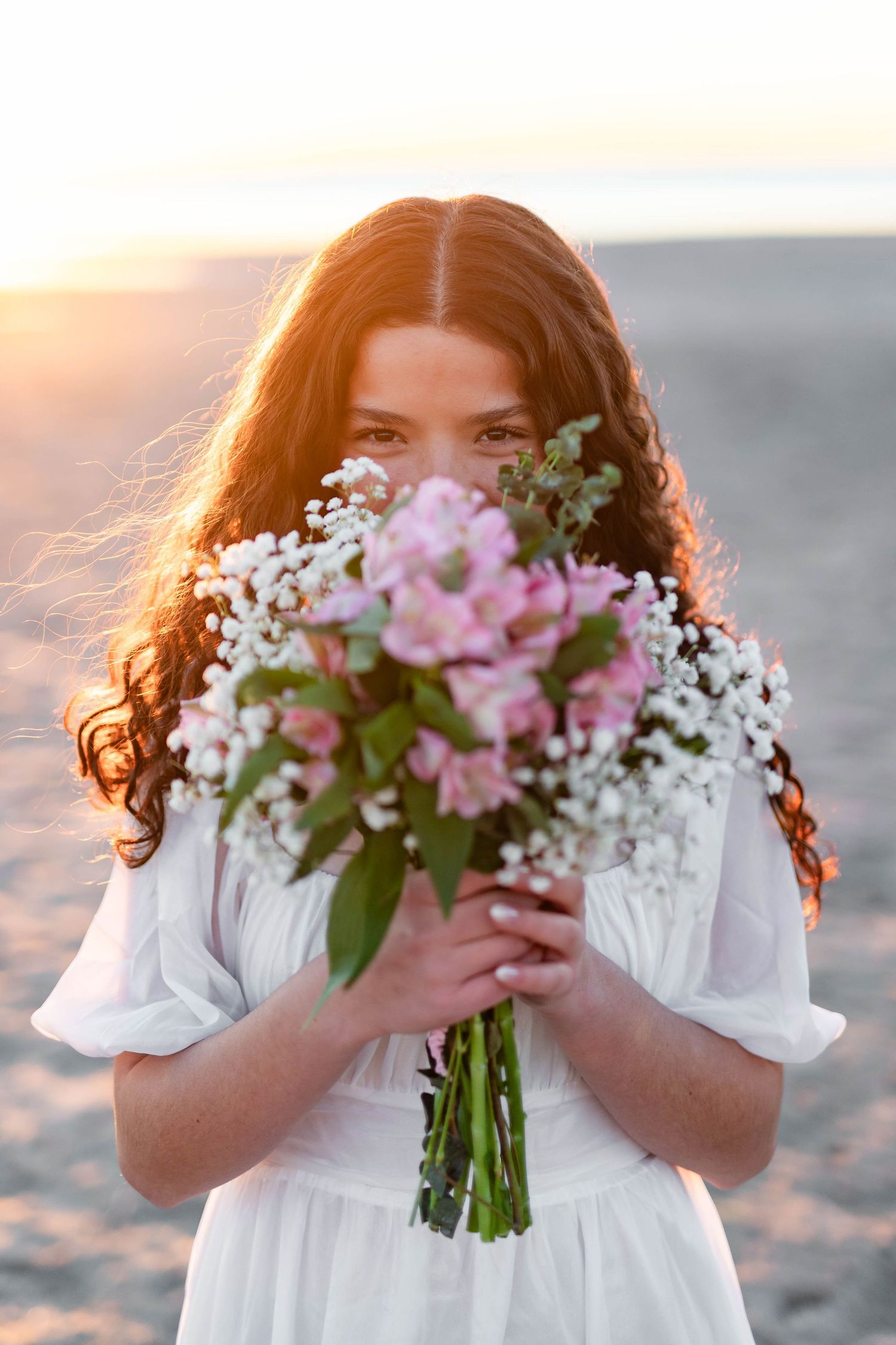Young woman with curly red hair in white dress holds pink and white flower bouquet at golden hour beach sunset.