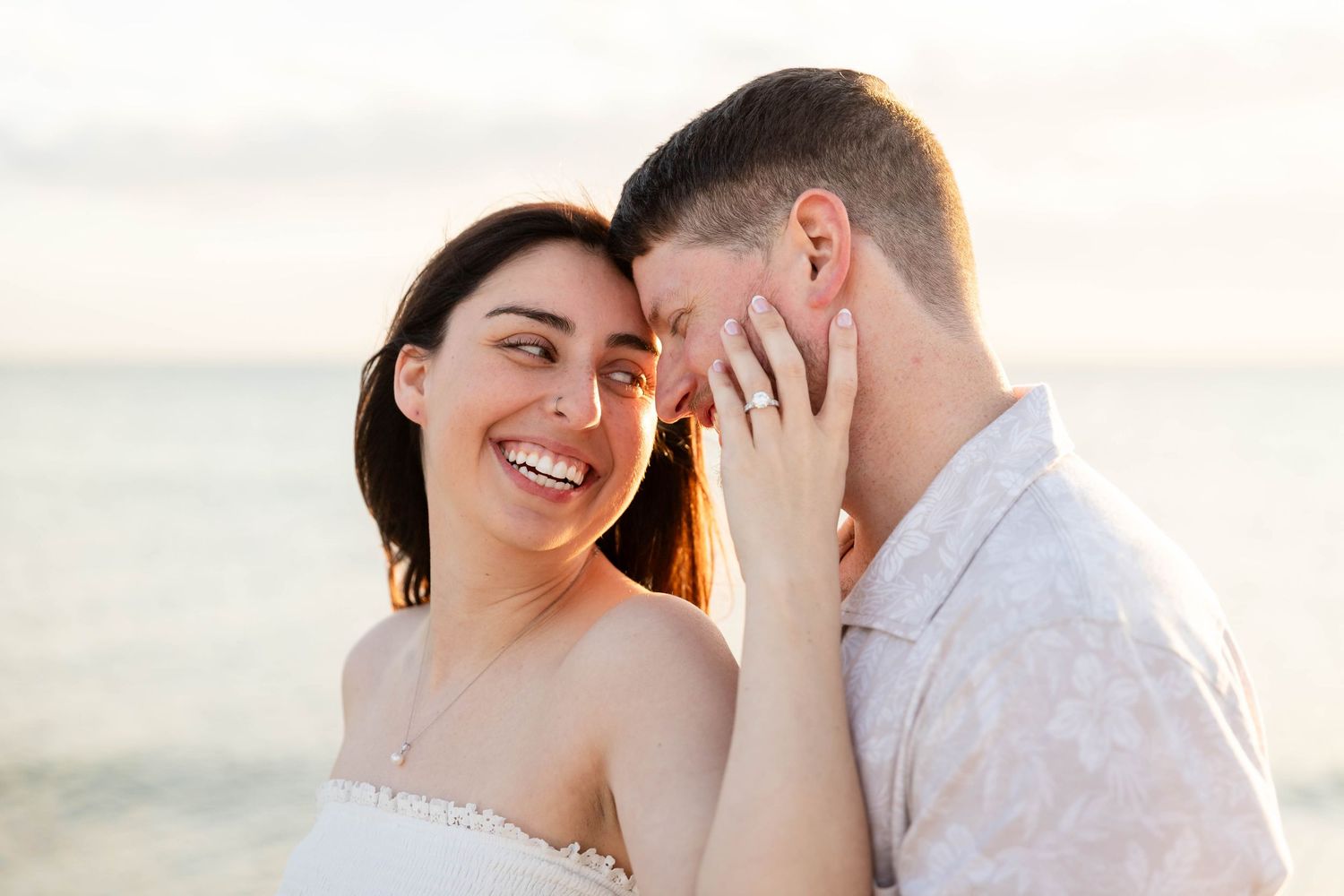 Smiling woman leaning close to her partner near the ocean during a golden sunset engagement photo at Honeymoon Island.