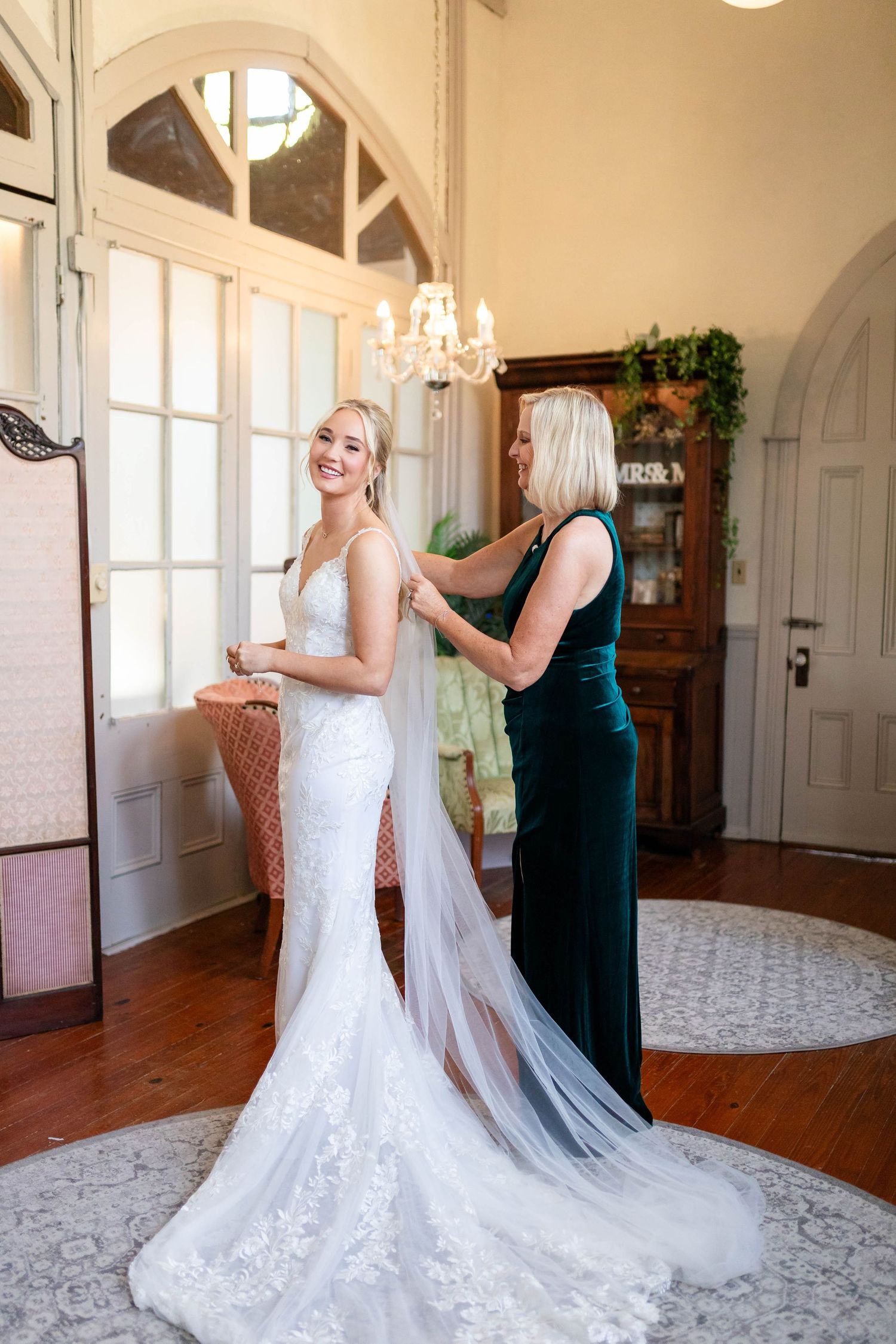 Smiling bride in lace mermaid gown having her veil fastened by a woman in a green velvet dress indoors in Dunedin.