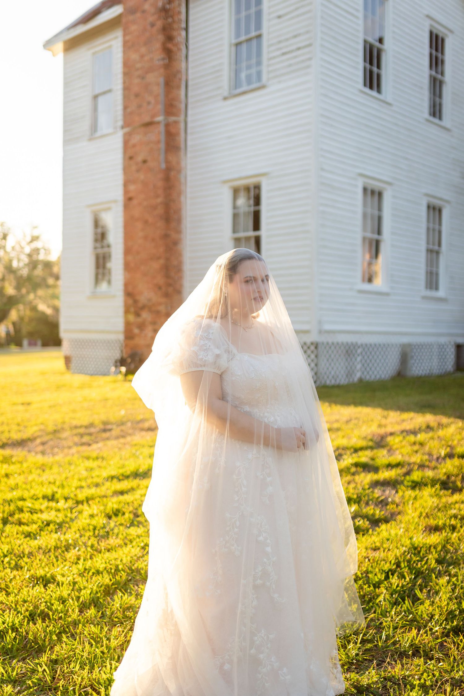 Bride in lace gown and flowing veil standing in golden sunlight before a white historic building.