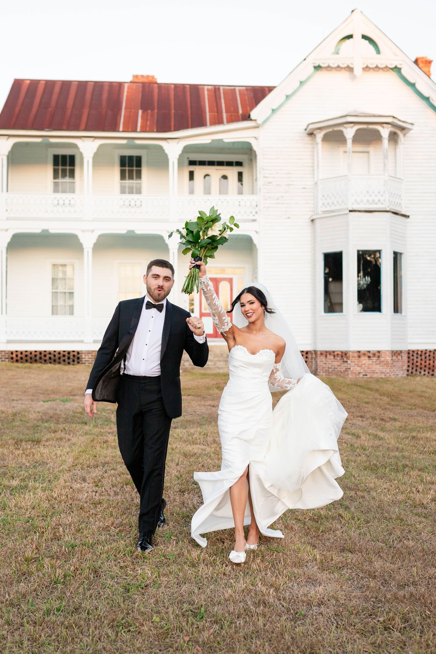 Joyful bride and groom walking hand in hand before a white Victorian wedding venue, bride raising bouquet.