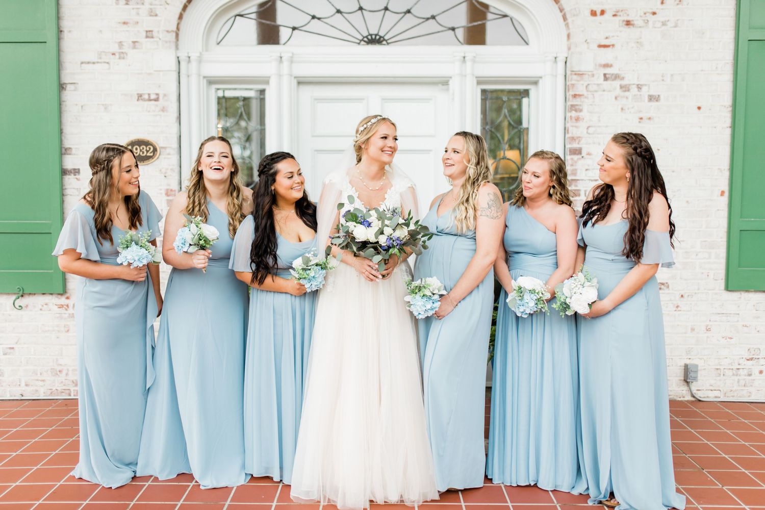 Bride and bridesmaids in powder blue dresses laughing together outside white brick venue entrance.