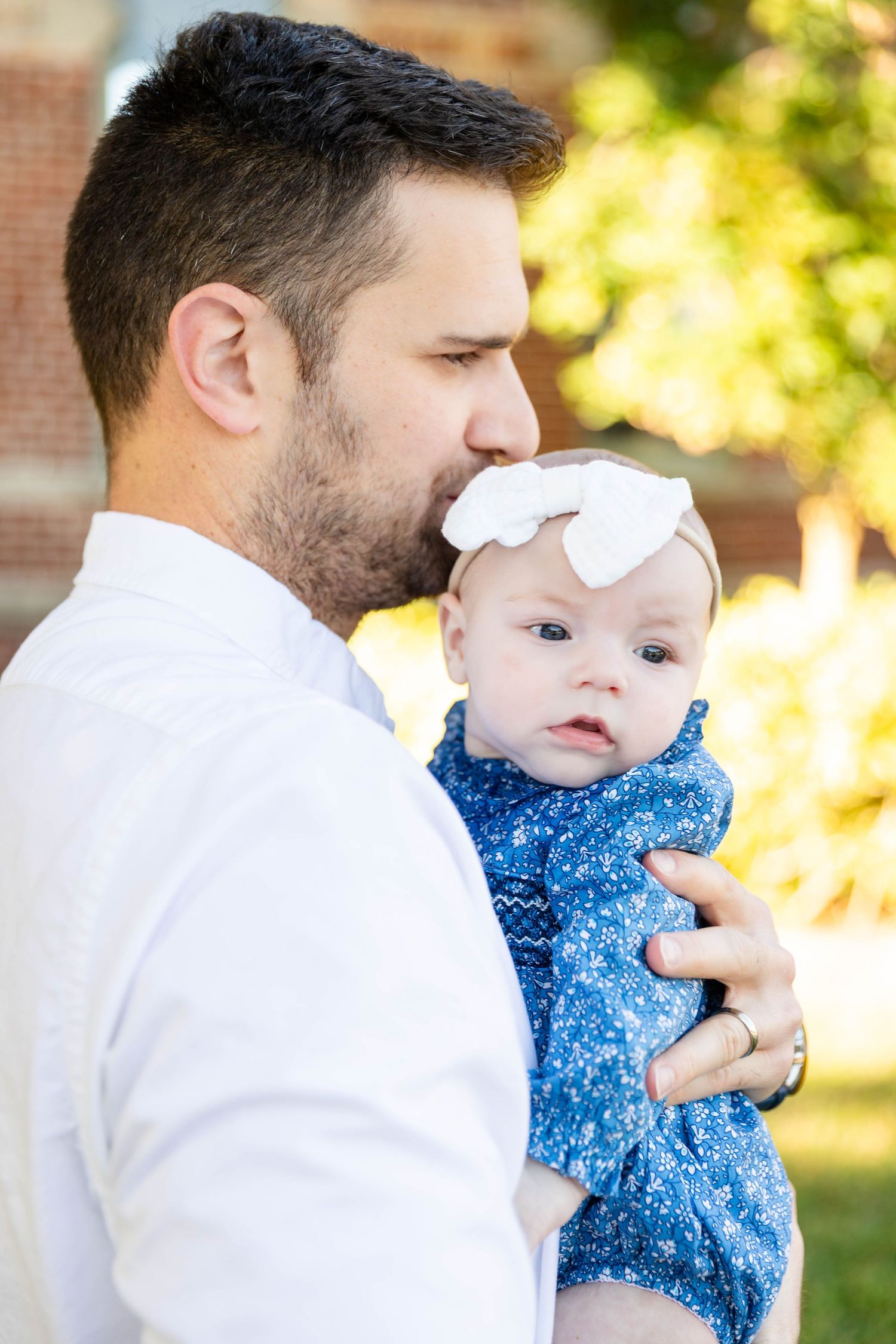 Father in white shirt gently kisses infant daughter wearing white bow headband outdoors at the University of Tampa.