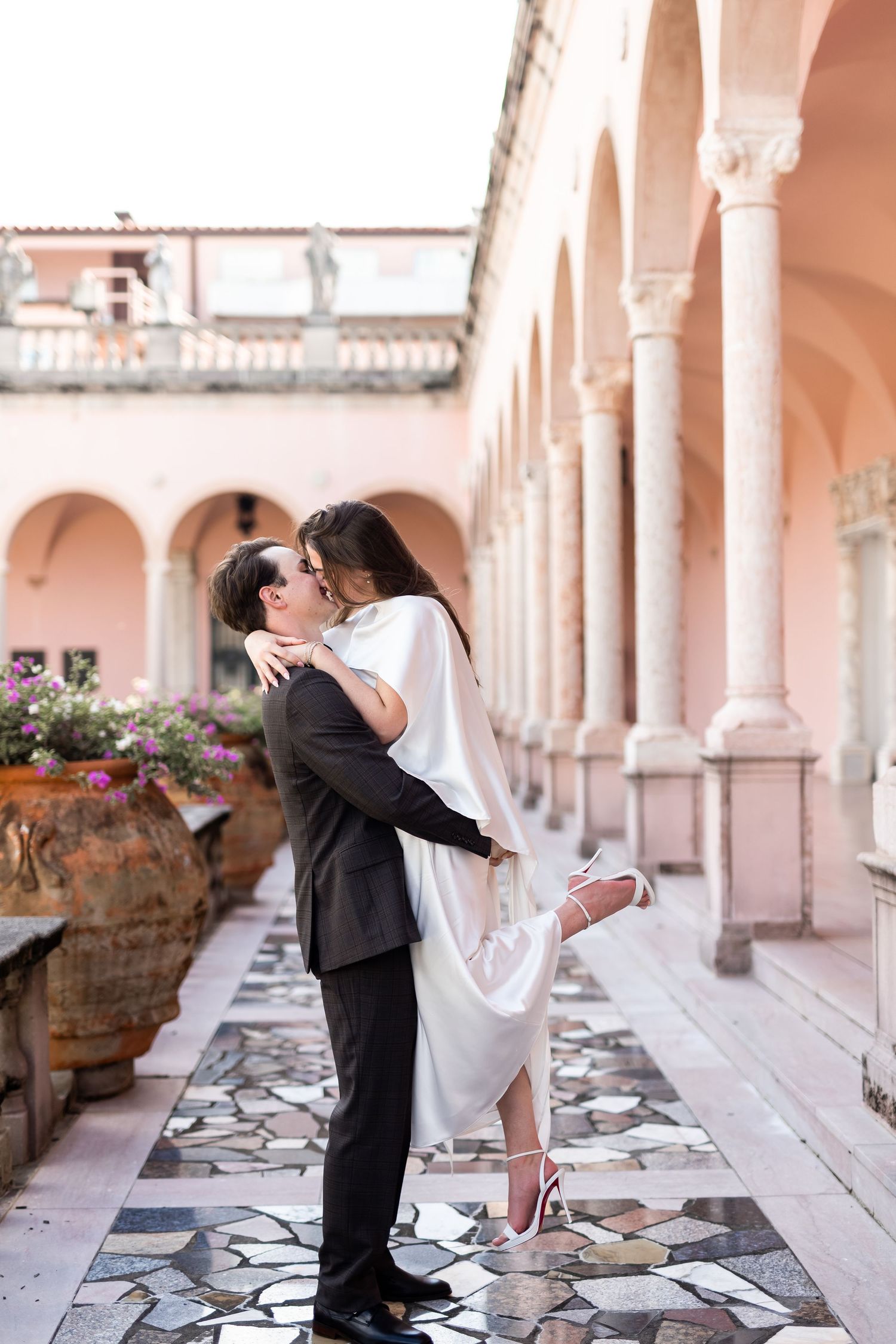 Bride and groom share romantic kiss in Italian courtyard colonnade with marble floors and pink walls at The Ringling Museum.