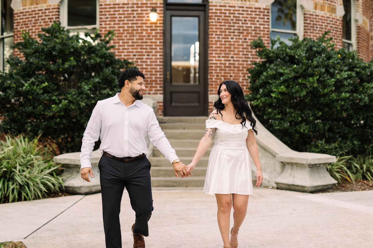 Couple holding hands and walking in front of a brick building, woman in white mini dress at the University of Tampa.