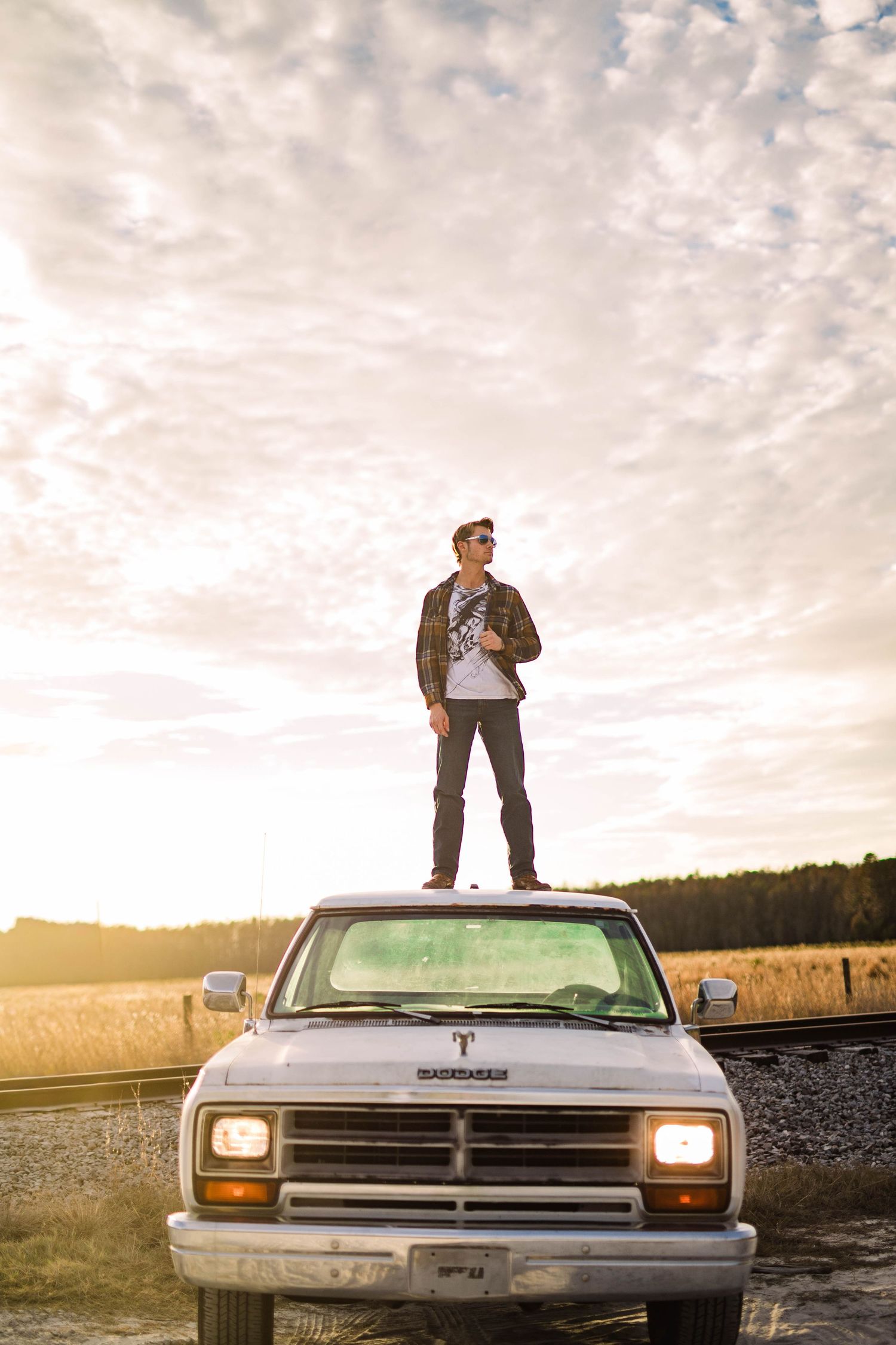 Young man standing on hood of white Dodge truck at sunset in rural field.
