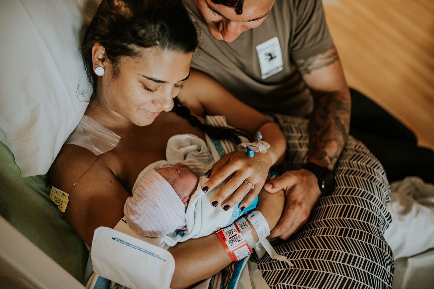 New mother holding newborn baby in hospital bed while father looks on lovingly.