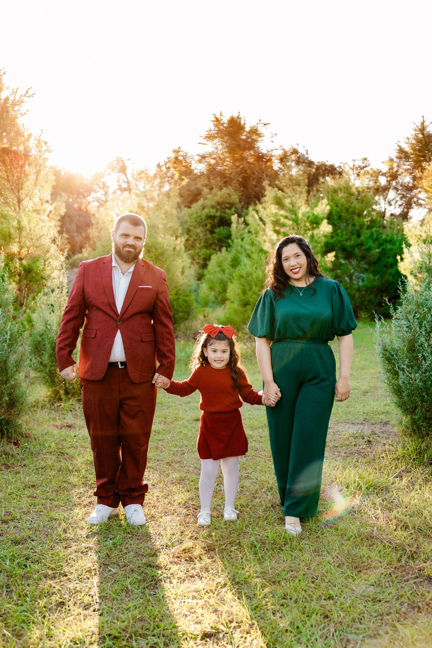 Family of three walks holding hands at Christmas tree farm, dressed in red and green holiday outfits.