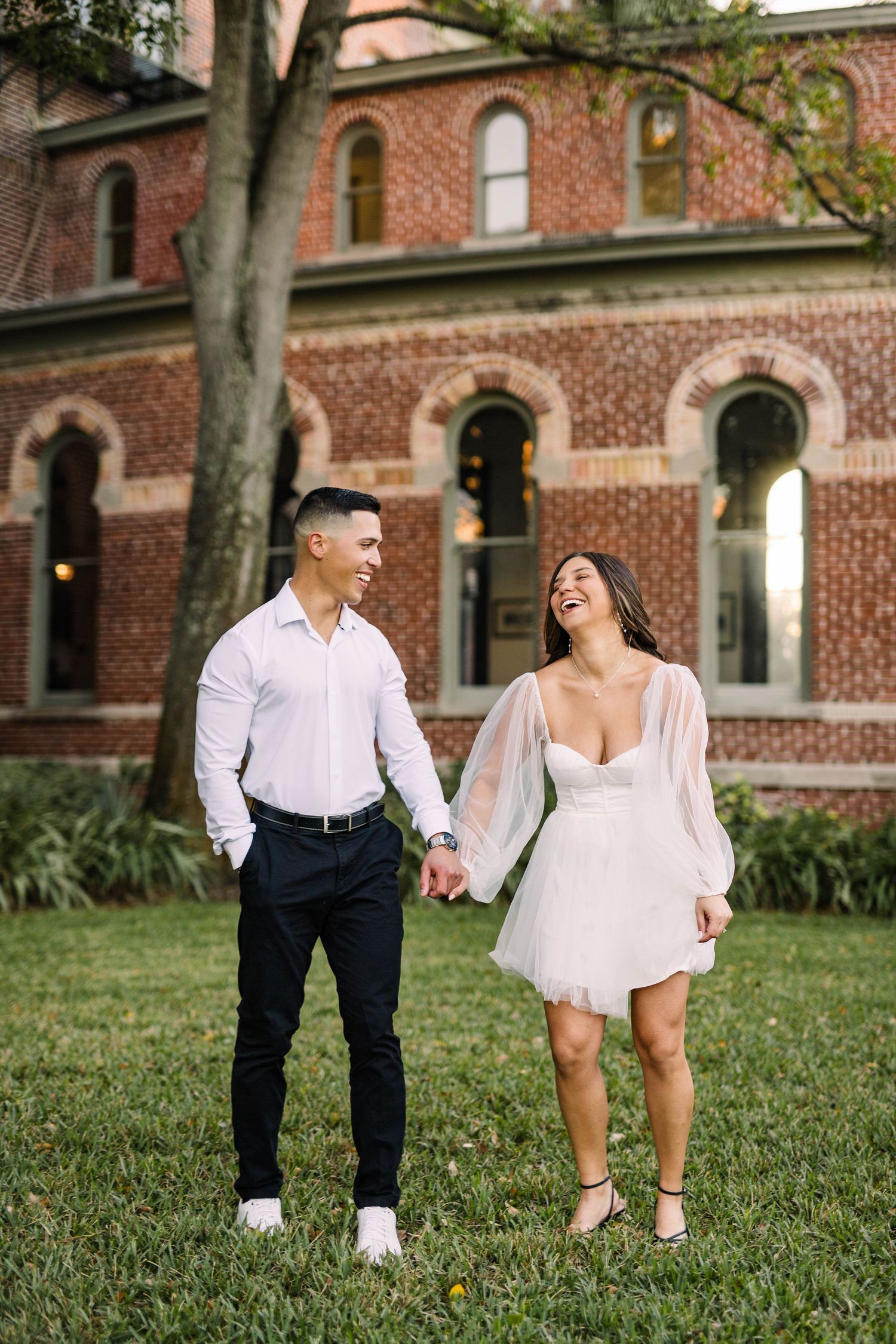 Happy couple holding hands and laughing in front of a historic red brick building on a green lawn at the University of Tampa.
