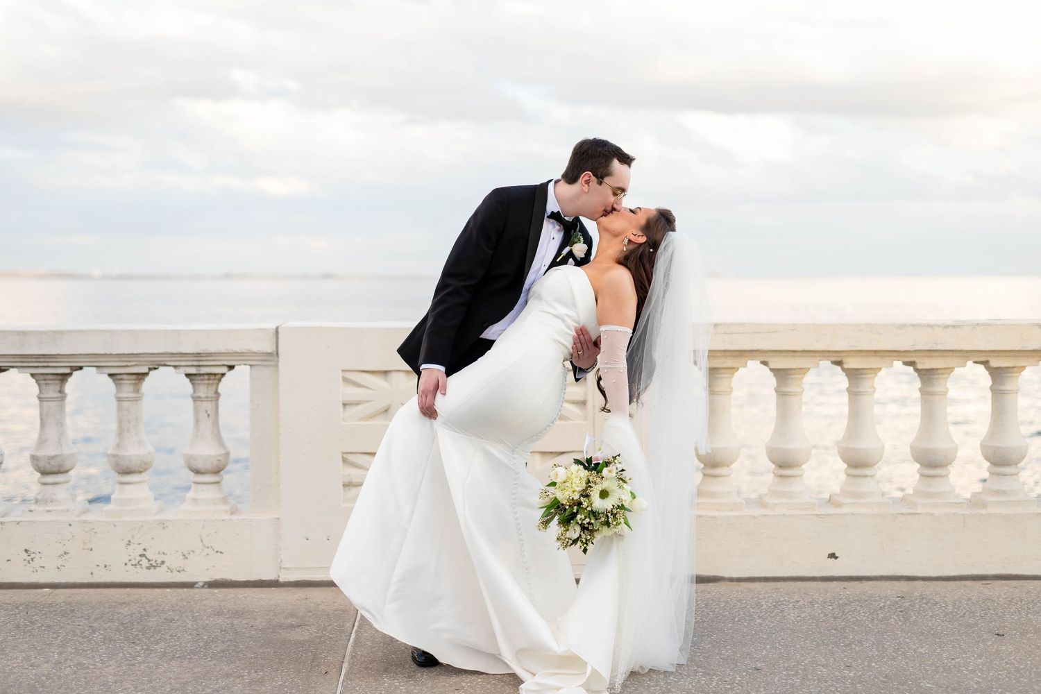 Bride and groom share a romantic kiss on a white balustrade terrace overlooking a cloudy sky on Boyshore Blvd in Tampa Florida.