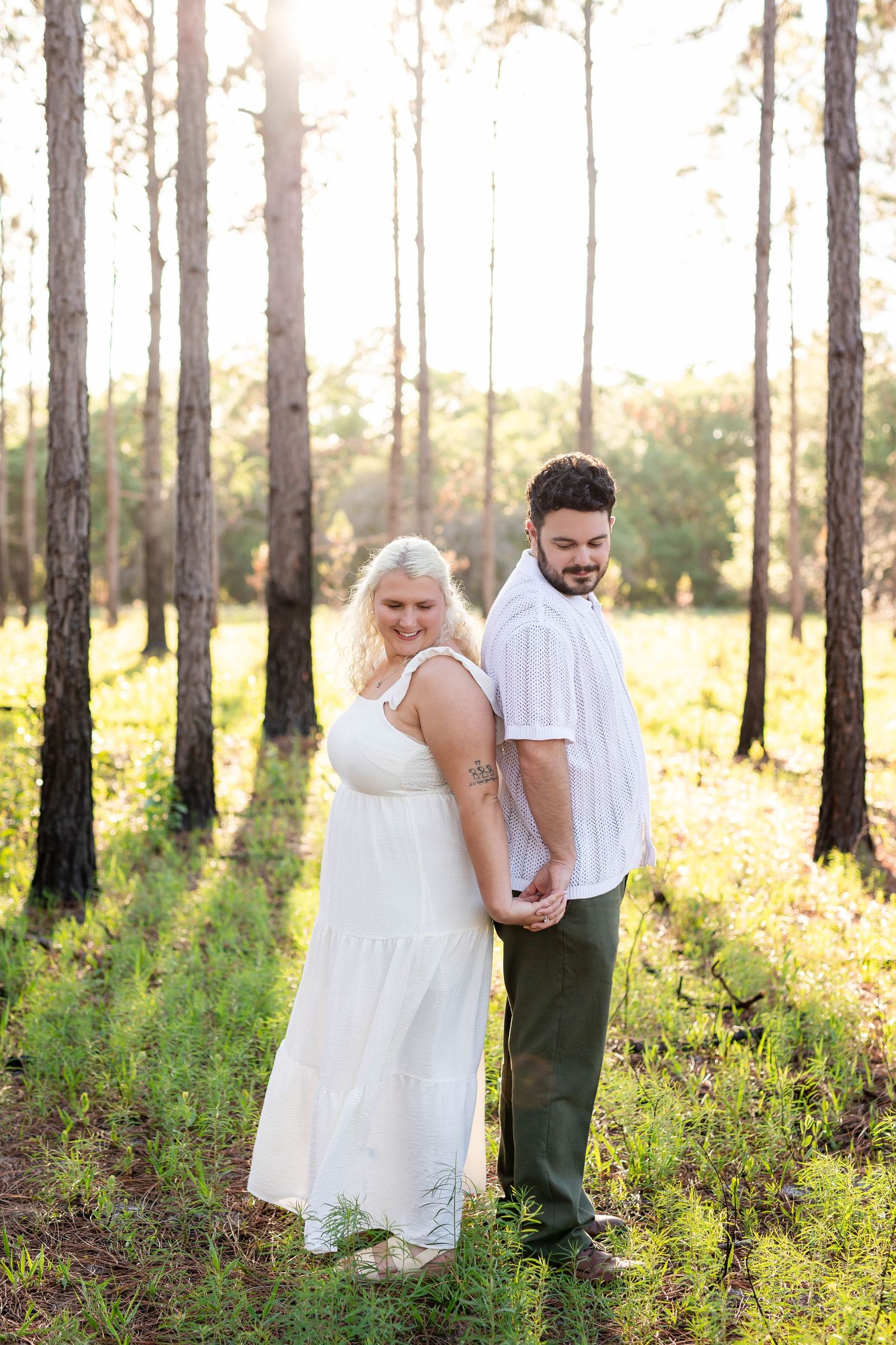 Couple holding hands back-to-back smiling in a sunlit pine forest during golden hour.