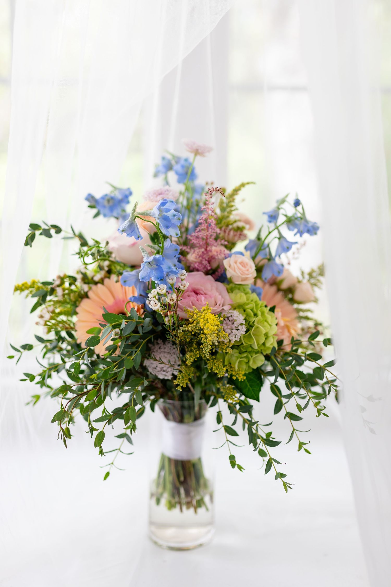Colorful wedding bouquet with pink peonies, blue delphinium, and greenery displayed in a glass vase.