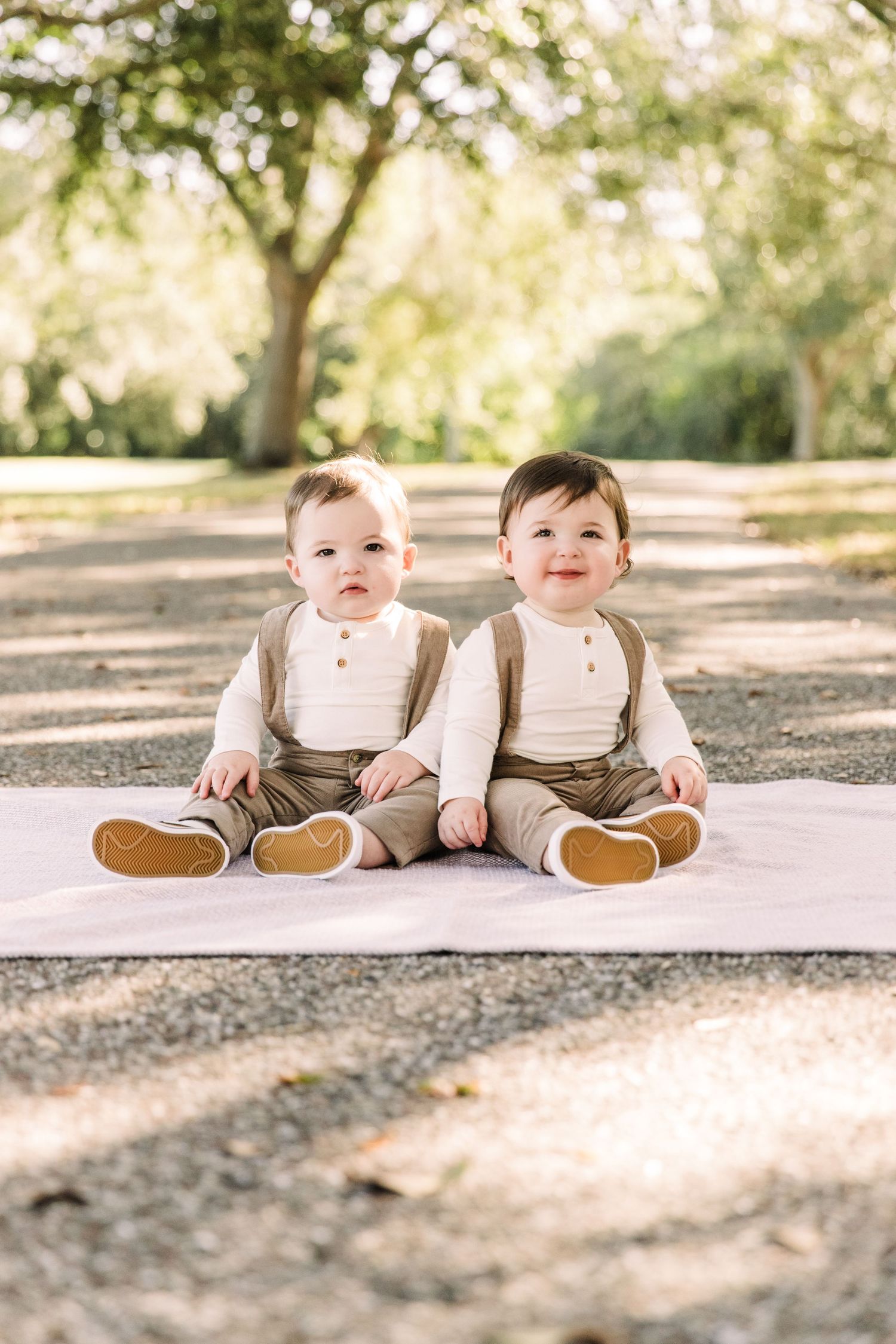 Adorable twin babies in matching brown suspender outfits sitting on blanket outdoors at Cypress Pointe Park.