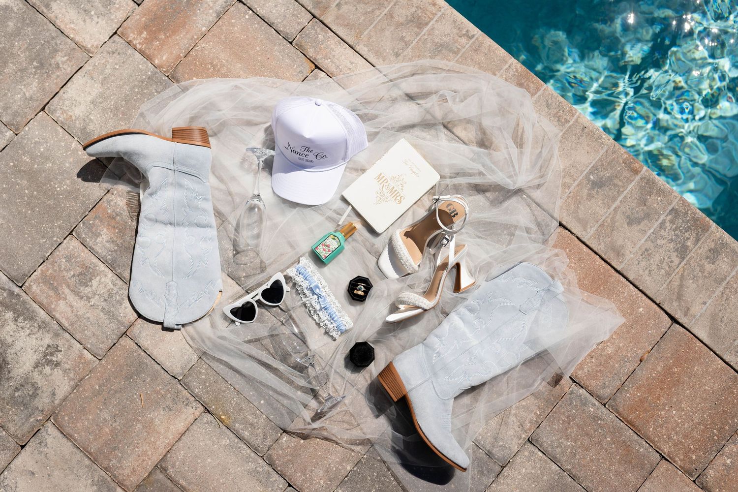 Flatlay of bridal accessories including boots, heels, sunglasses, and a veil arranged on brick pavers by a pool in Dunedin.