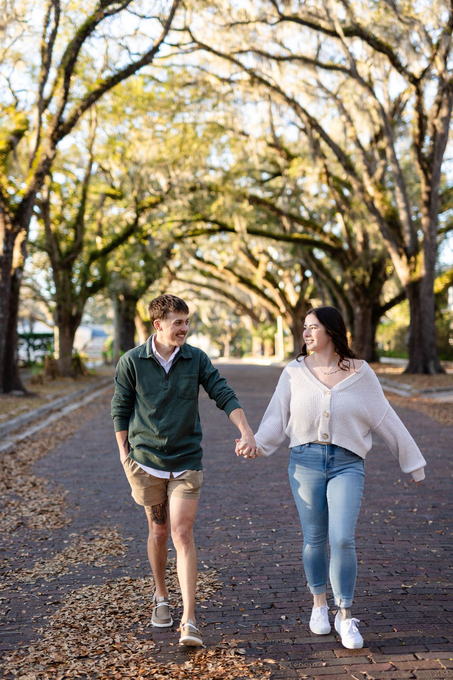 Happy couple holding hands and walking along a brick path lined with beautiful oak trees.