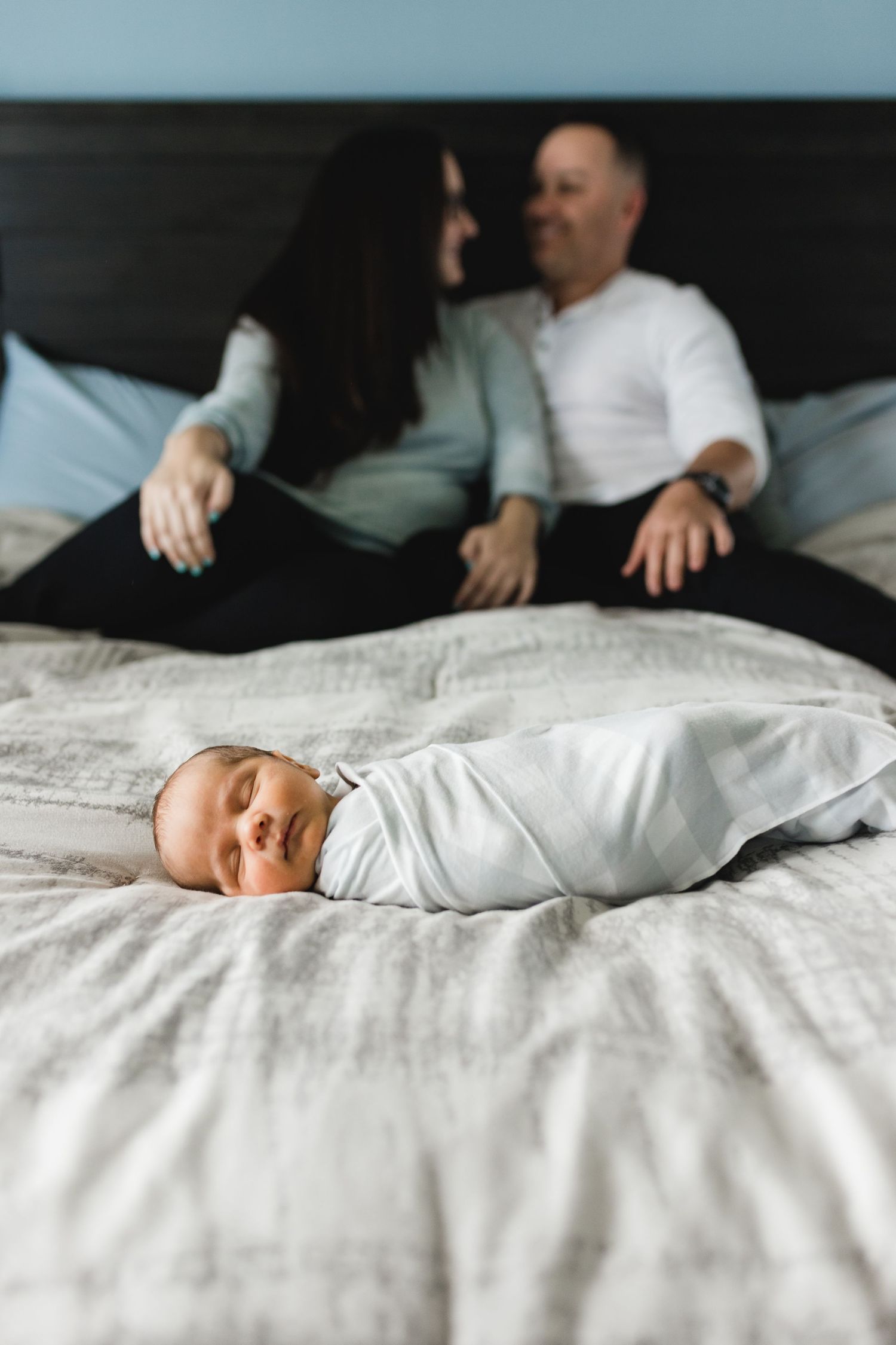 Newborn baby swaddled in white lying on bed with parents sitting together in the background.