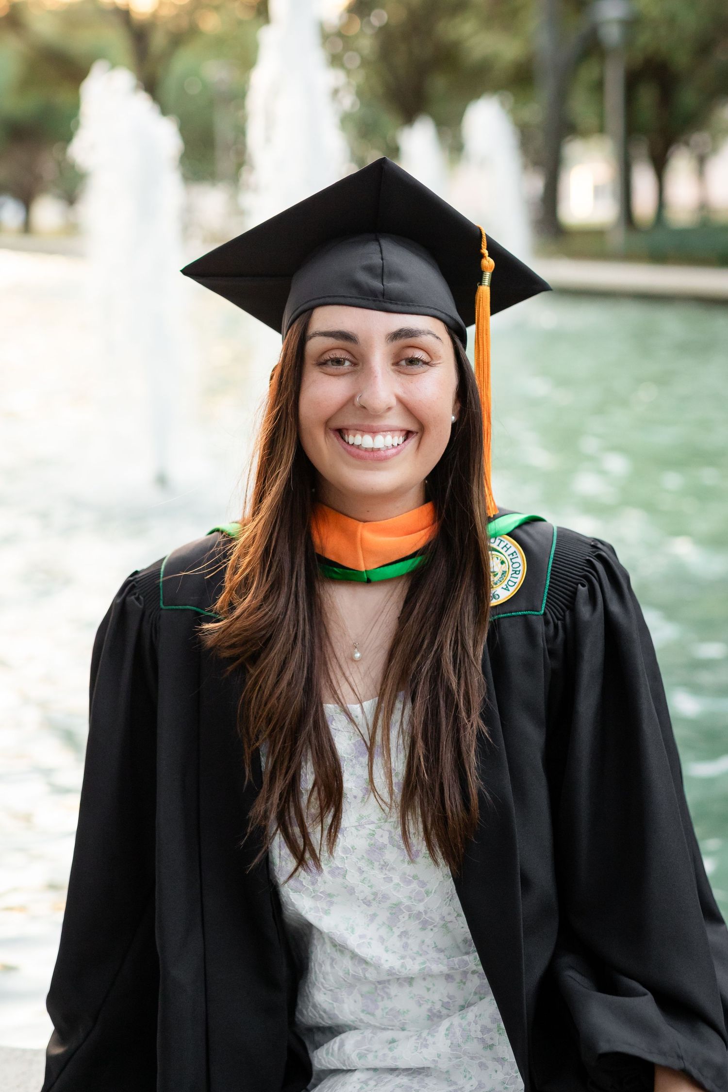 Smiling female graduate in black cap and gown with orange and green hood posing near a fountain at the University of South Florida.
