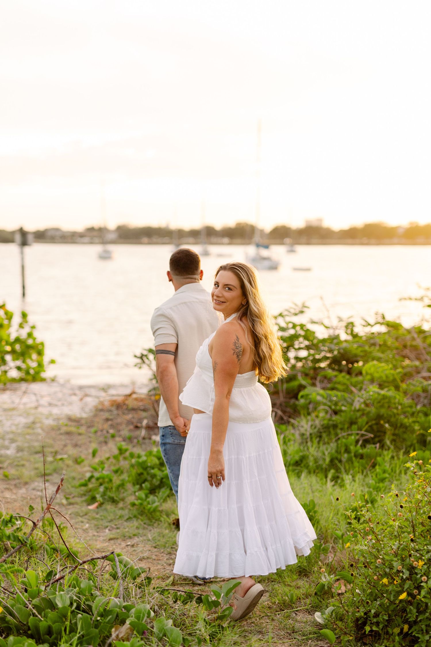 Woman in white dress looks back smiling while holding hands with partner near golden sunset waterfront at Davis Island.