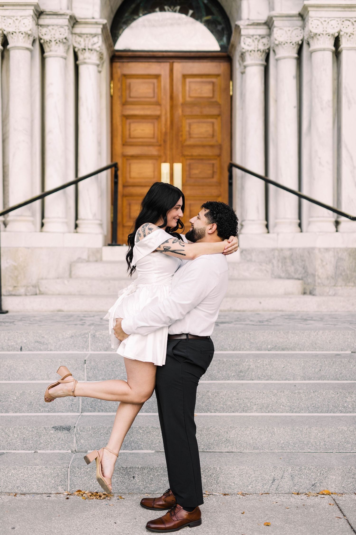 Man lifting woman in white dress on marble church steps, both laughing in a joyful embrace in Tampa.