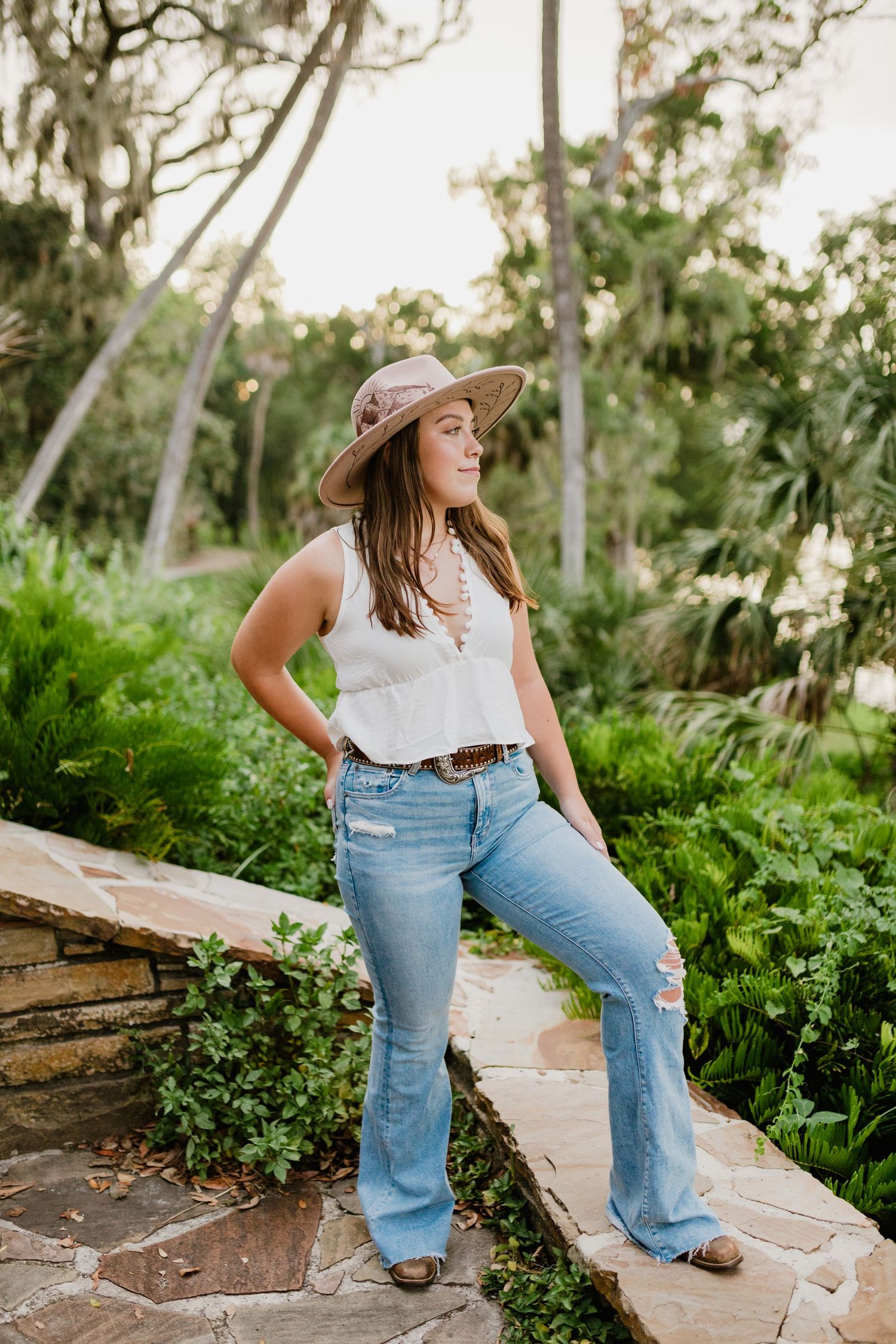 Young woman in white top, flare jeans, and wide-brim hat poses on stone path surrounded by lush greenery.