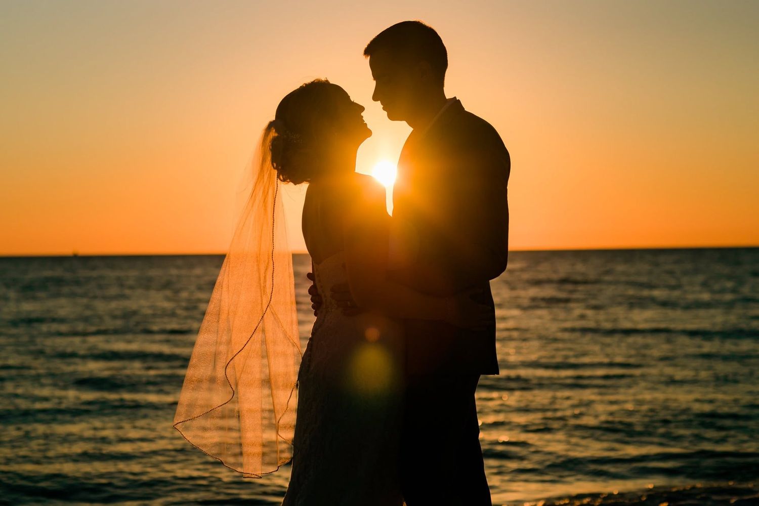 Romantic silhouette of bride and groom nearly kissing against stunning ocean sunset backdrop.