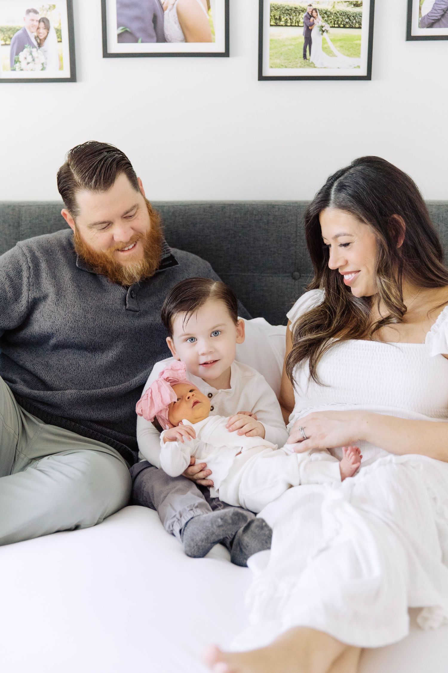 Happy family with red-bearded father, brunette mother, toddler, and newborn baby sitting on couch.