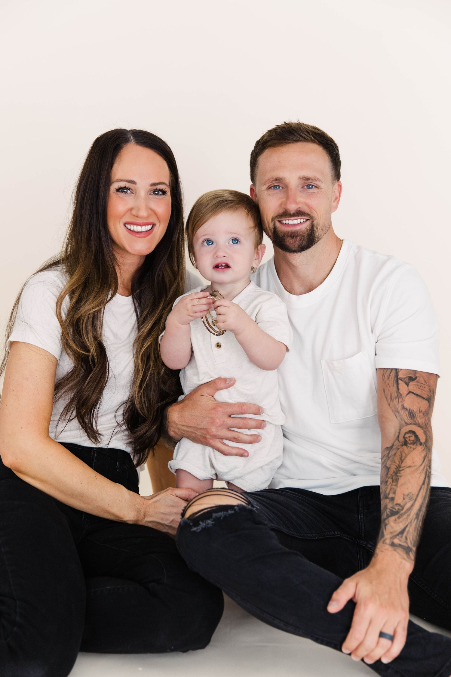 Happy family with tattooed dad, brunette mom, and blue-eyed baby in white studio.