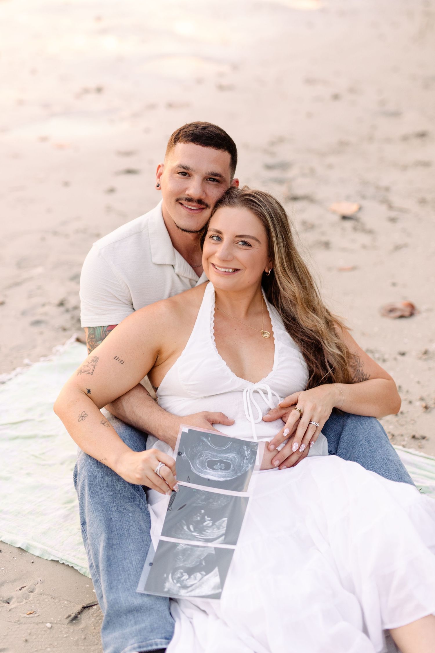 Happy couple sits on beach holding ultrasound images, celebrating pregnancy announcement at sunset at Davis Island.