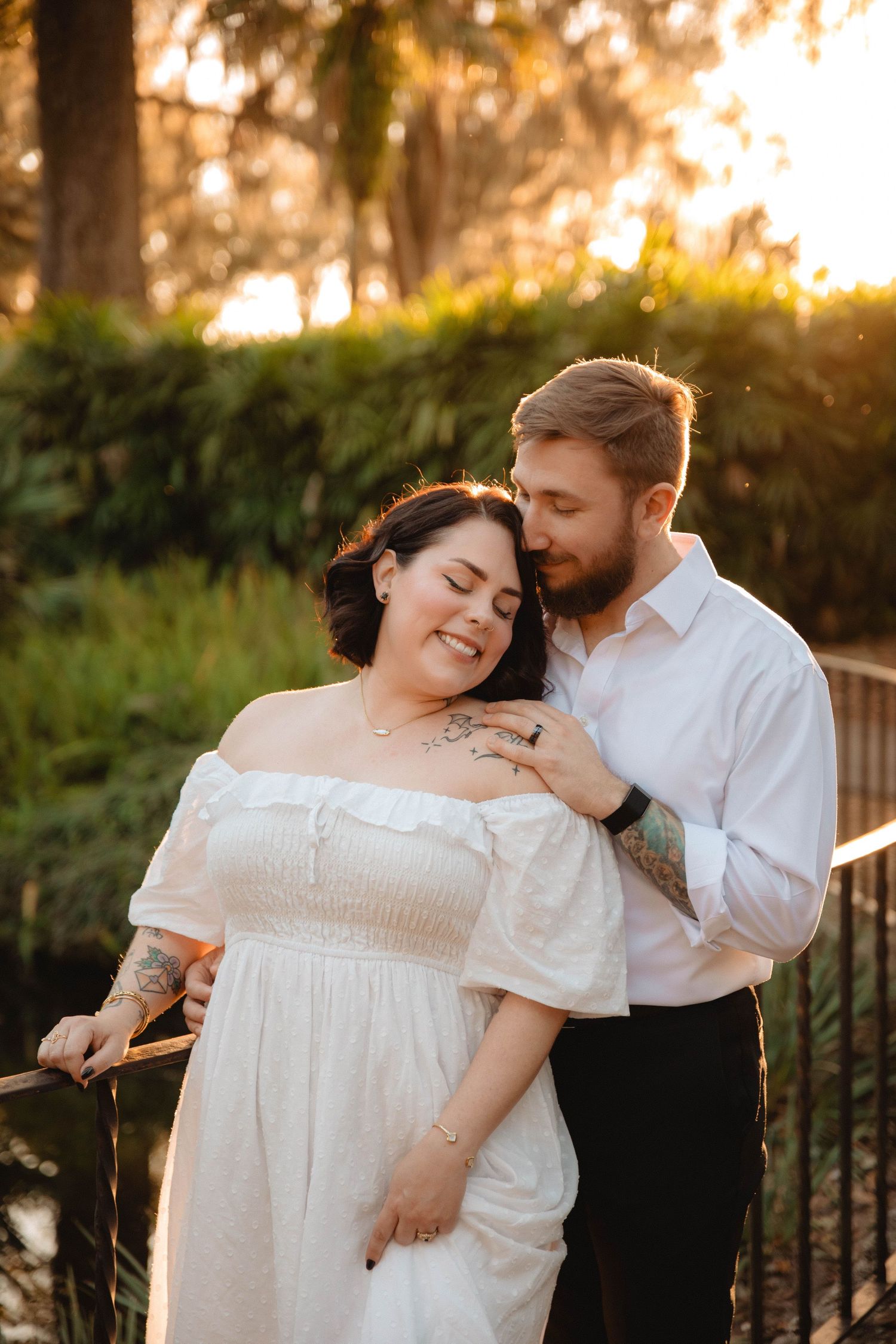 Couple sharing a tender embrace on a bridge at sunset, woman in white off-shoulder dress smiling at Bok Tower Gardens, Lake Wales.
