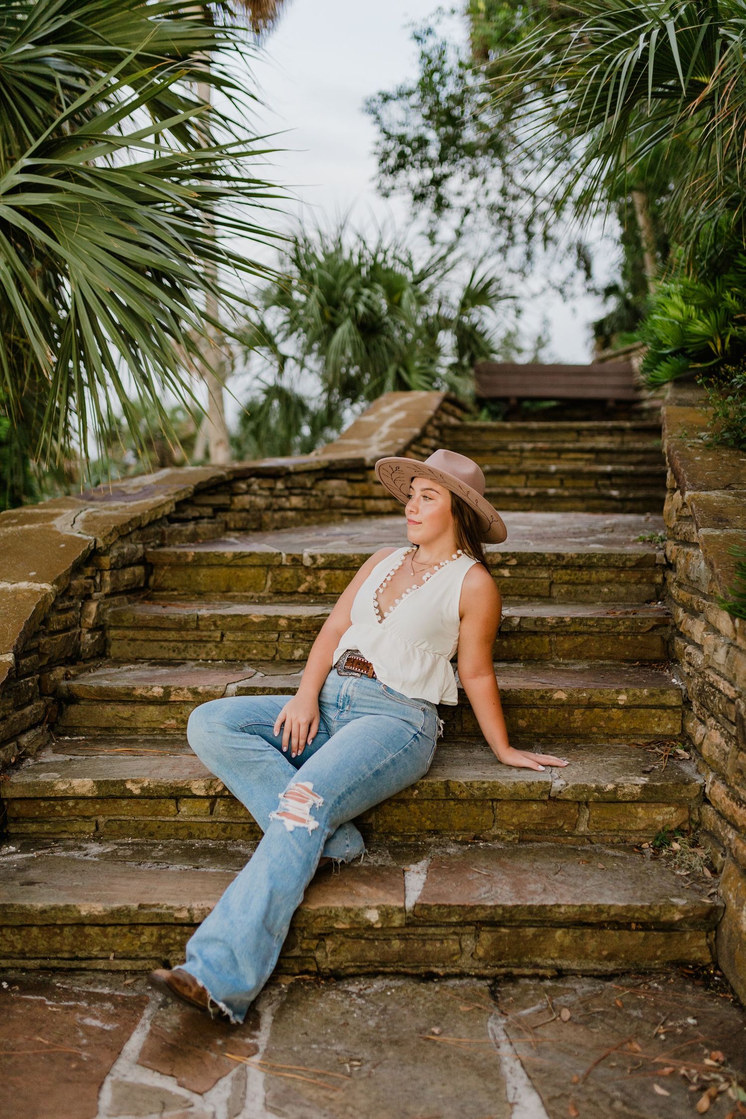Young woman in cowboy hat and ripped jeans sitting on stone steps surrounded by tropical palm trees.