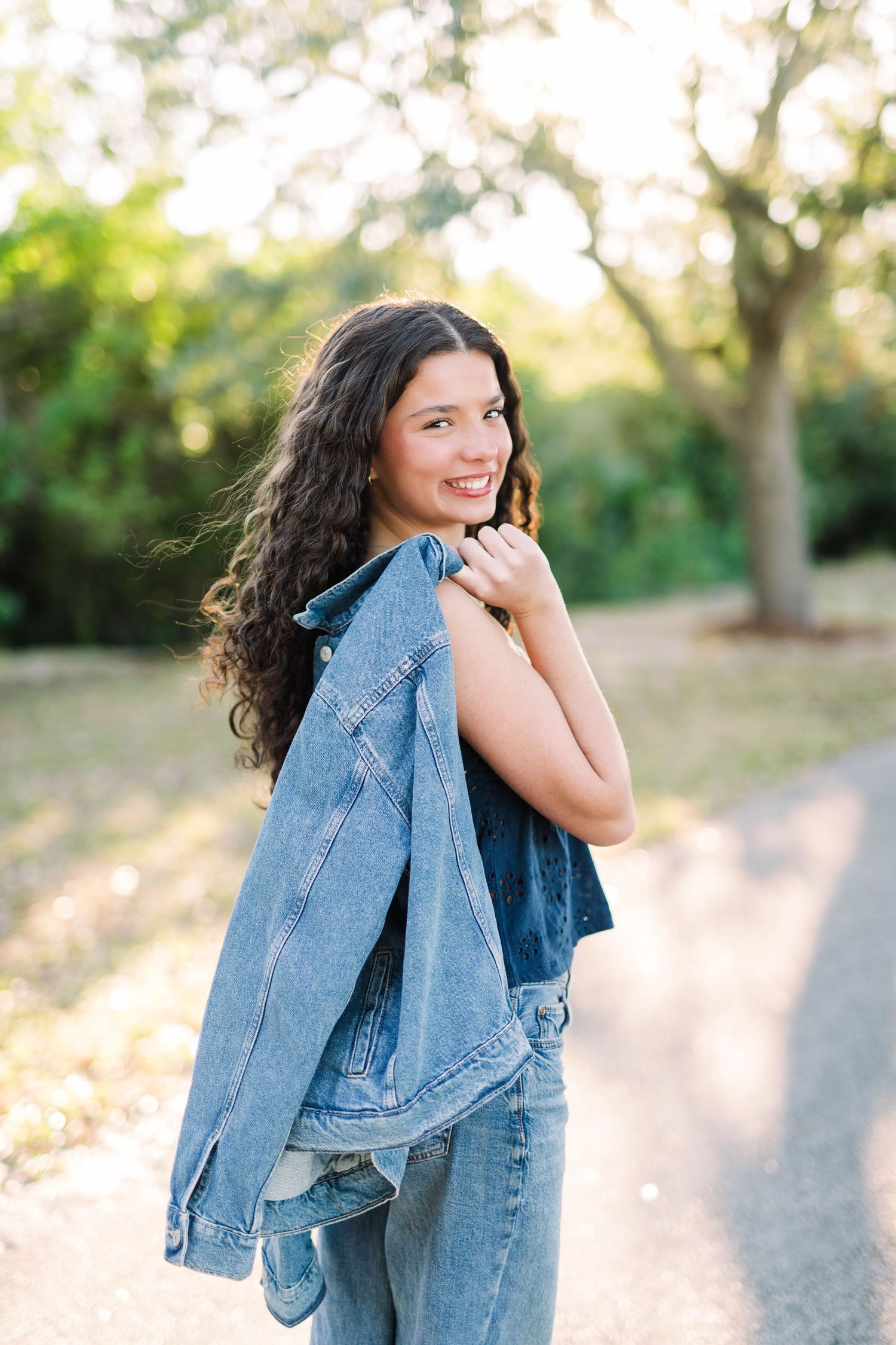 Teen girl in denim jacket and jeans smiling over shoulder on sunlit park path with curly hair.