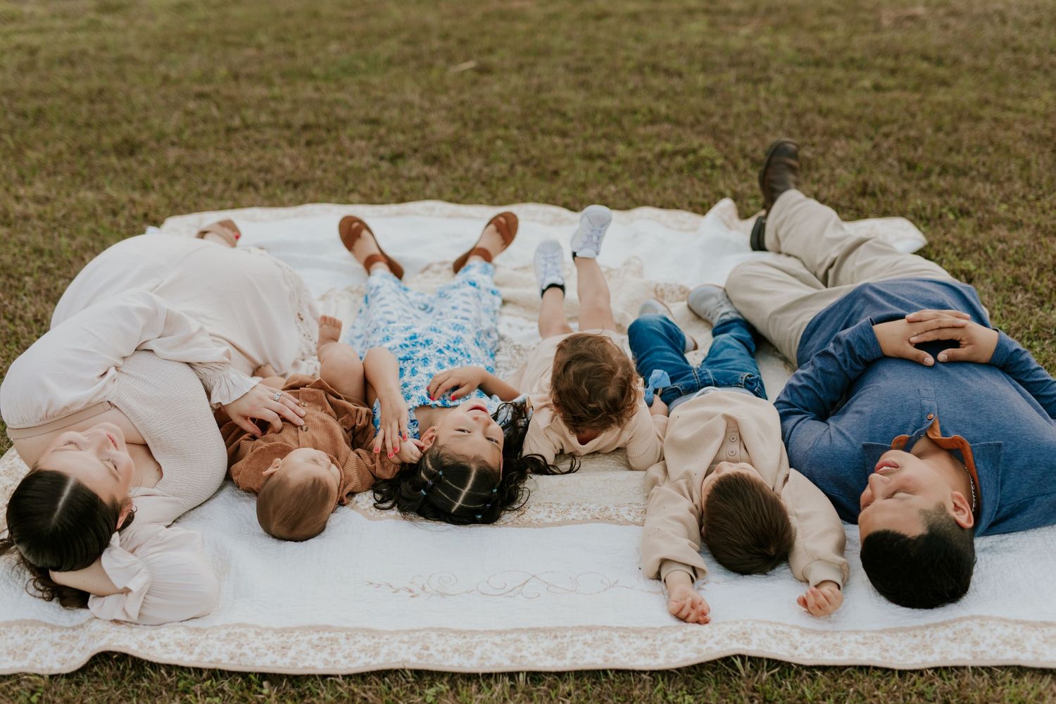 Family laying together on a white blanket in the grass, smiling and enjoying a sunny outdoor day.