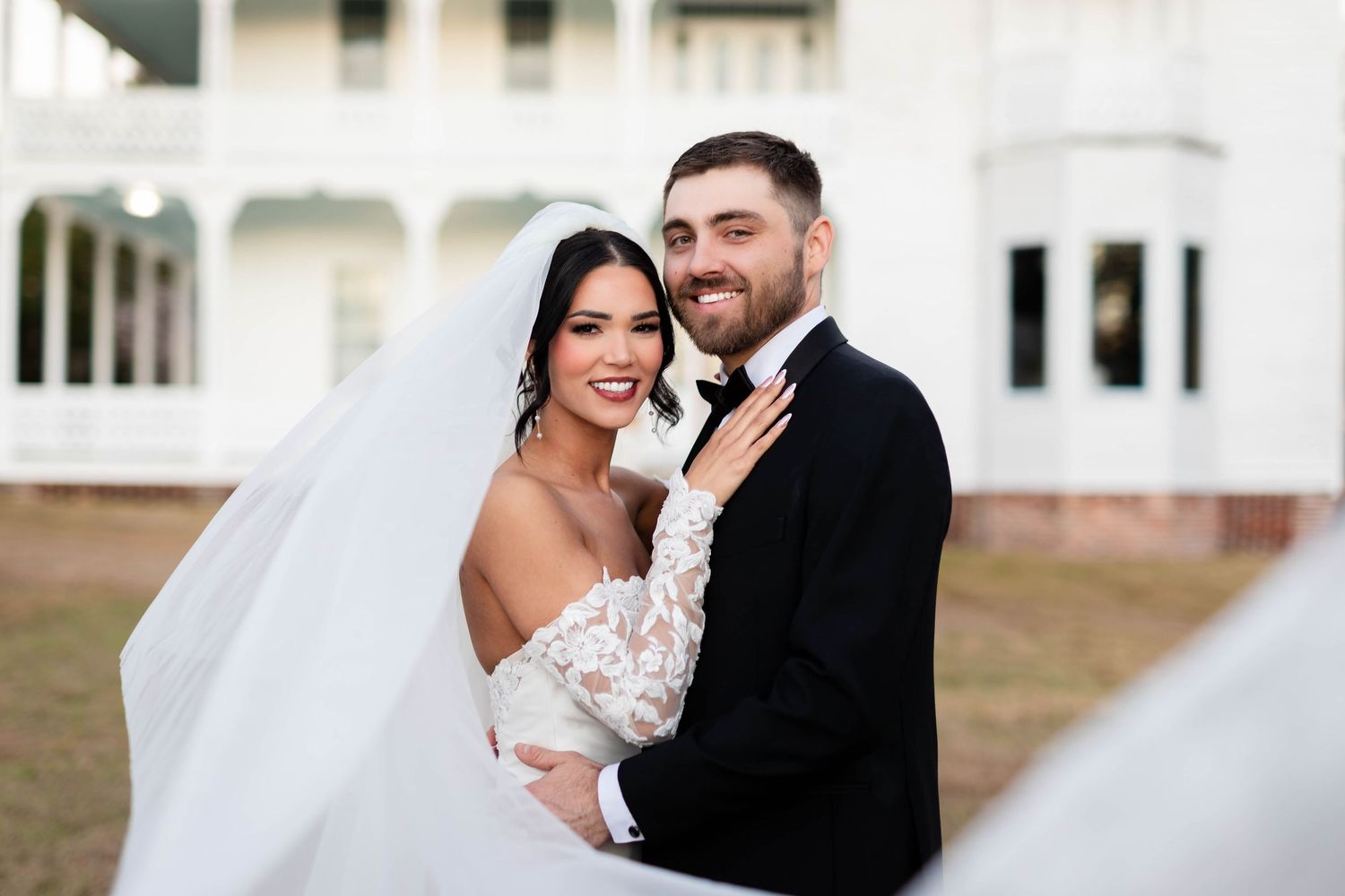 Bride in lace-sleeved gown embraces smiling groom in black tuxedo outside a white historic building.