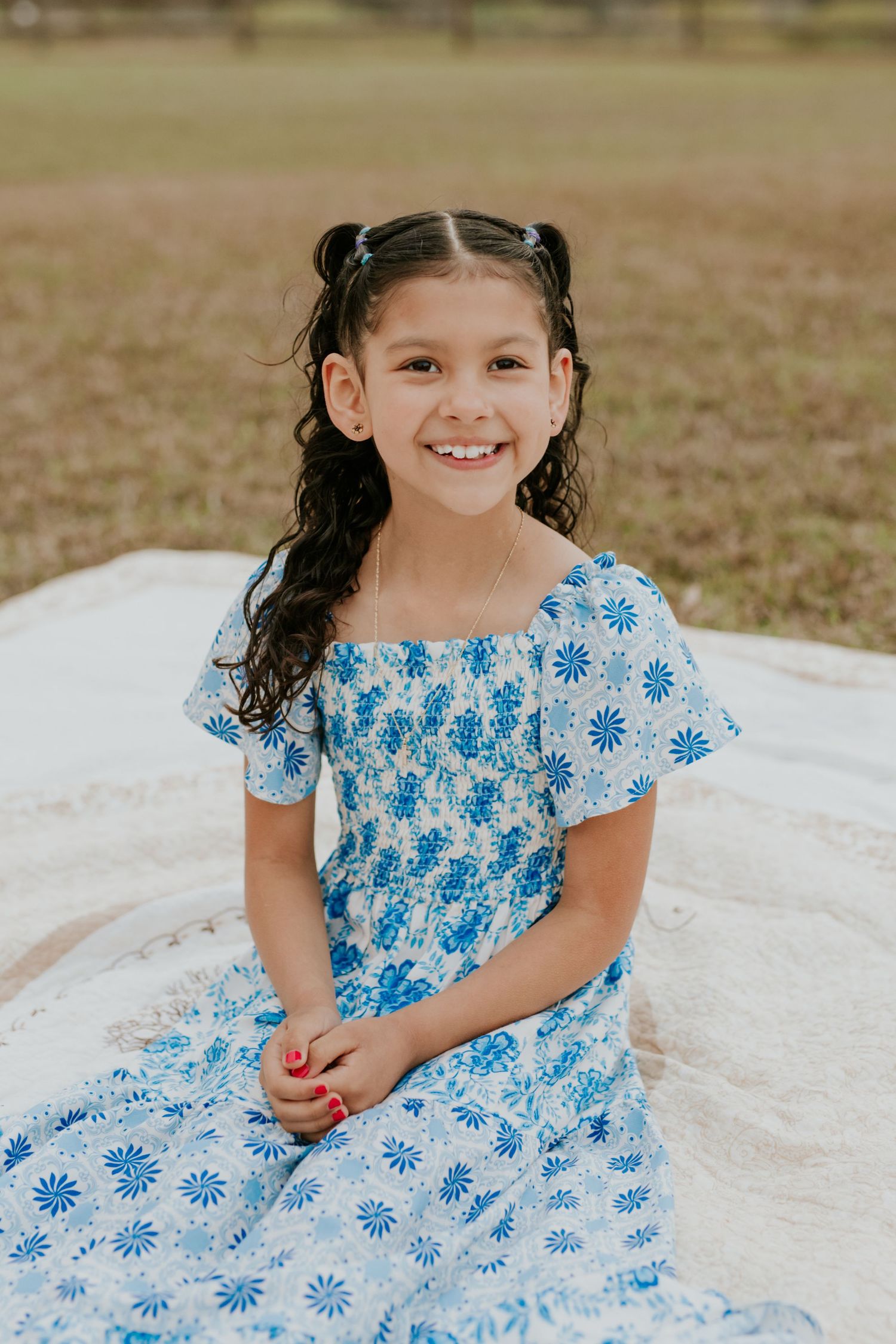 Young girl in blue floral dress with pigtails smiling while sitting on blanket.