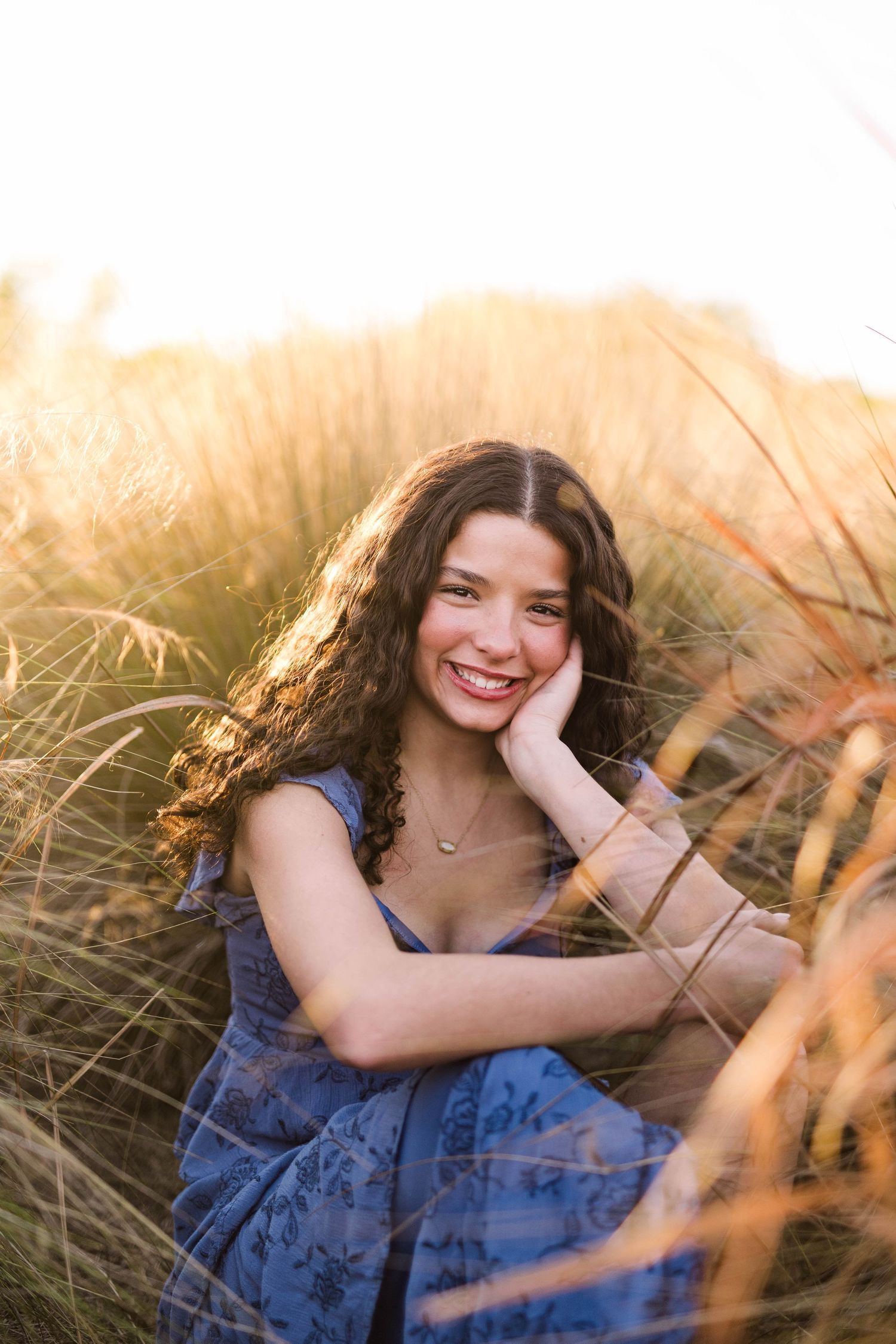 Young woman in blue dress sitting in golden grass field at sunset, smiling with hand on cheek.